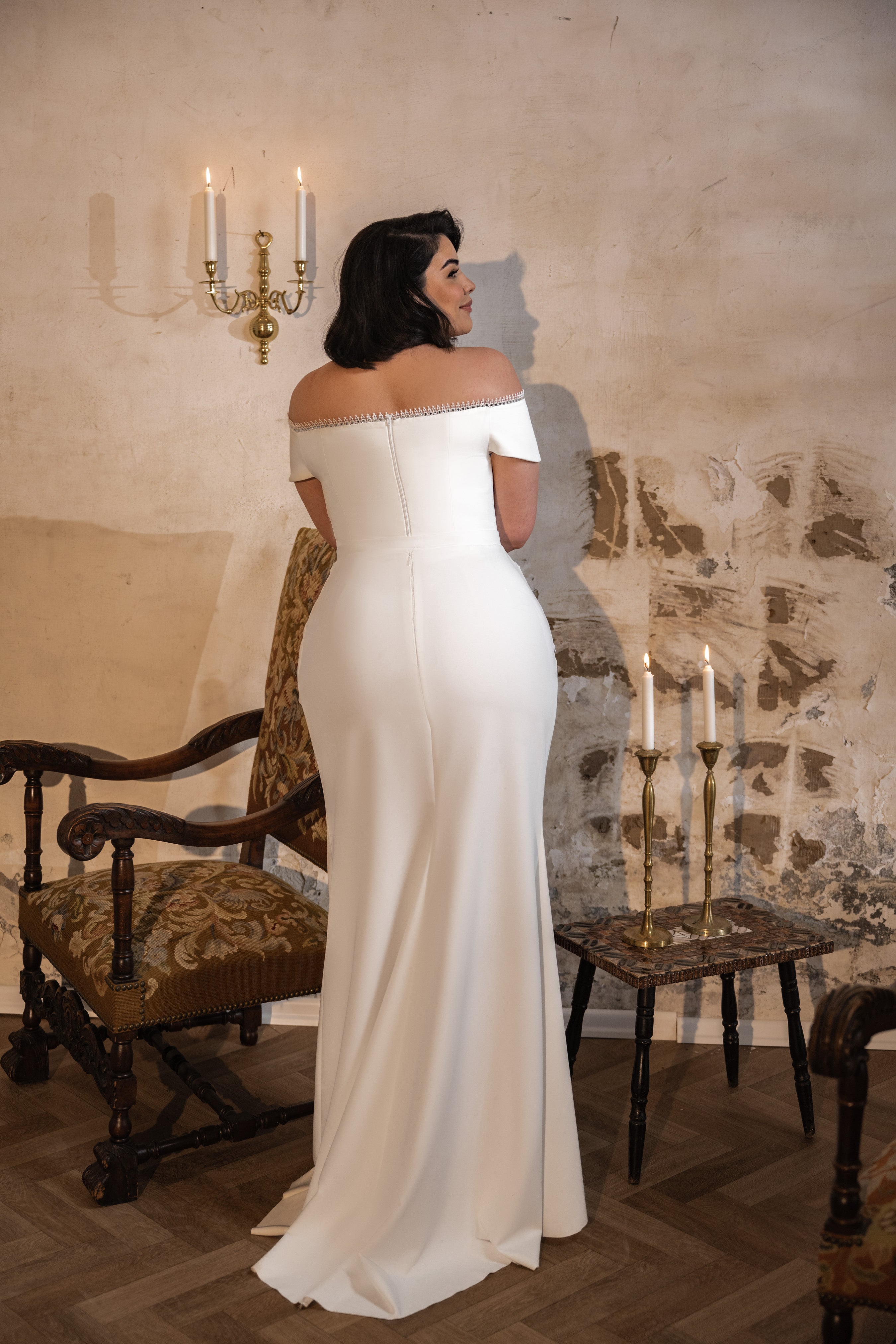 אישה לבושה בשמלת Annabelle מקולקציית Bridal Plus 2023/24 עומדת עם גבה למצלמה, מול תפאורה וינטג'ית עם קיר מרקם, שני פמוטים מוארים, כיסא מעוטר ושולחן קטן.
