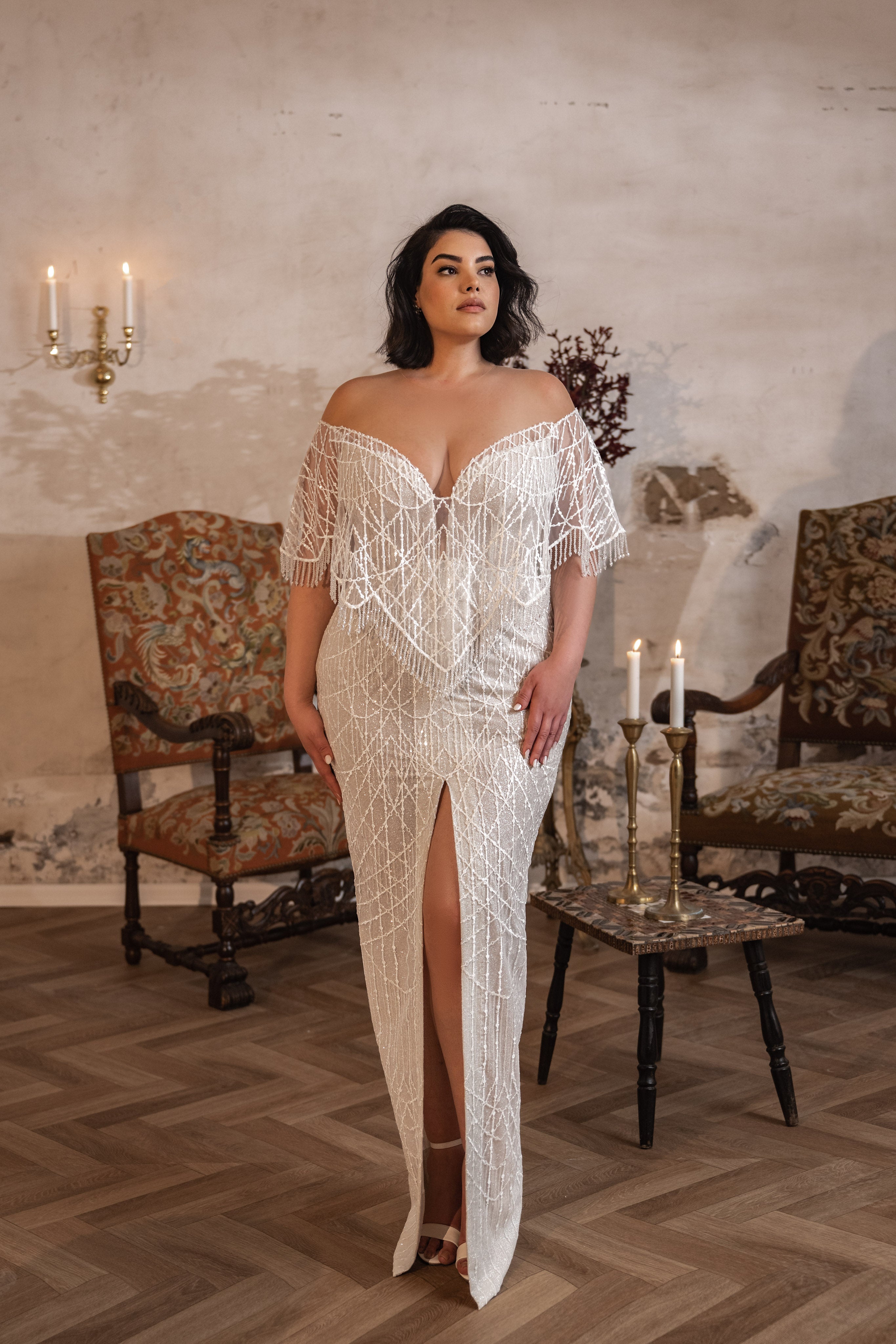 אישה לובשת את קולקציית Bridal Plus 2023/24 - Diana: שמלת כלה אלגנטית, עם כתפיים חשופות, מעוטרת בקריסטלים, עומדת בתוך חדר כפרי מואר באור רך, בין כורסאות מעוטרות ונרות.