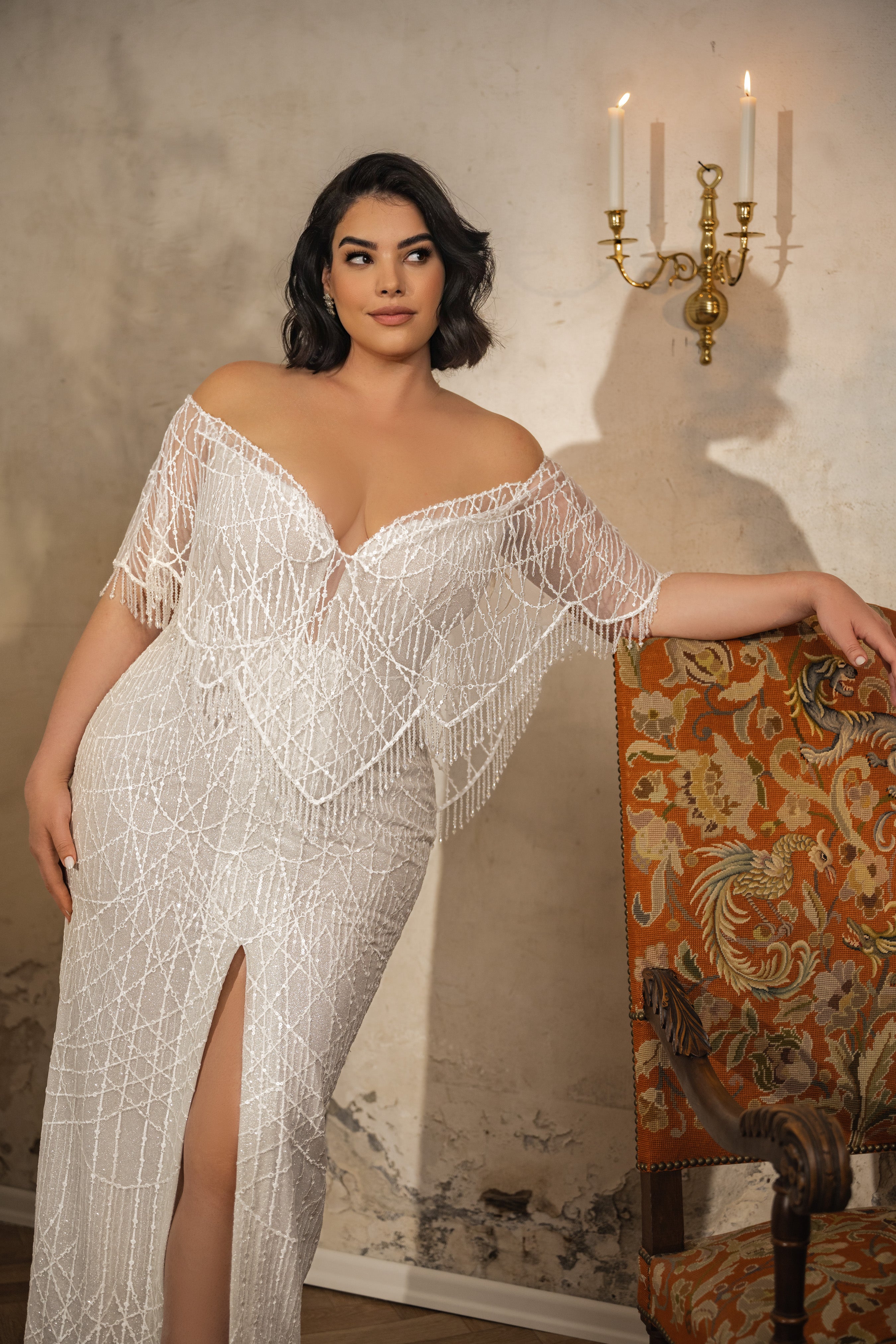 אישה לבושה בשמלת כלה מקולקציית Bridal Plus 2023/24 - Diana, שמלת כלה לבנה וזוהרת עם כתפיים חשופות, מעוטרת בחרוזים ובקישוטי קריסטל, מצטלמת ליד כיסא מעוטר ופמוט קיר בחדר בסגנון וינטג'.