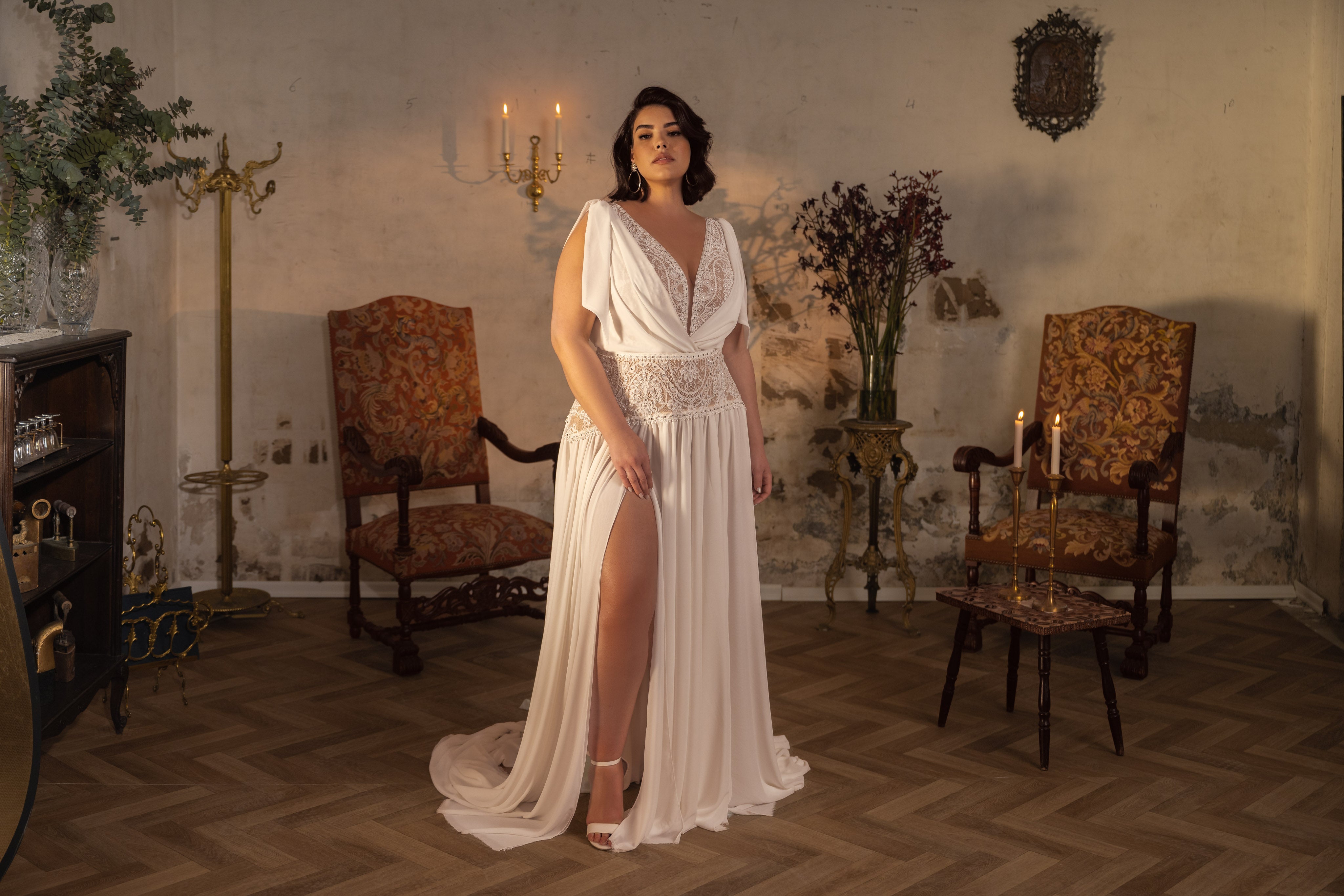 אישה מקולקציית Bridal Plus 2023/24 - שמלת כלה משיפון לבן של קריסטן עם חתך גבוה עד הירך עומדת בביטחון בחדר בסגנון וינטג' עם כיסאות מעוטרים, עיצוב פרחוני ואור נרות, המעוררים אווירה רומנטית וקלאסית.