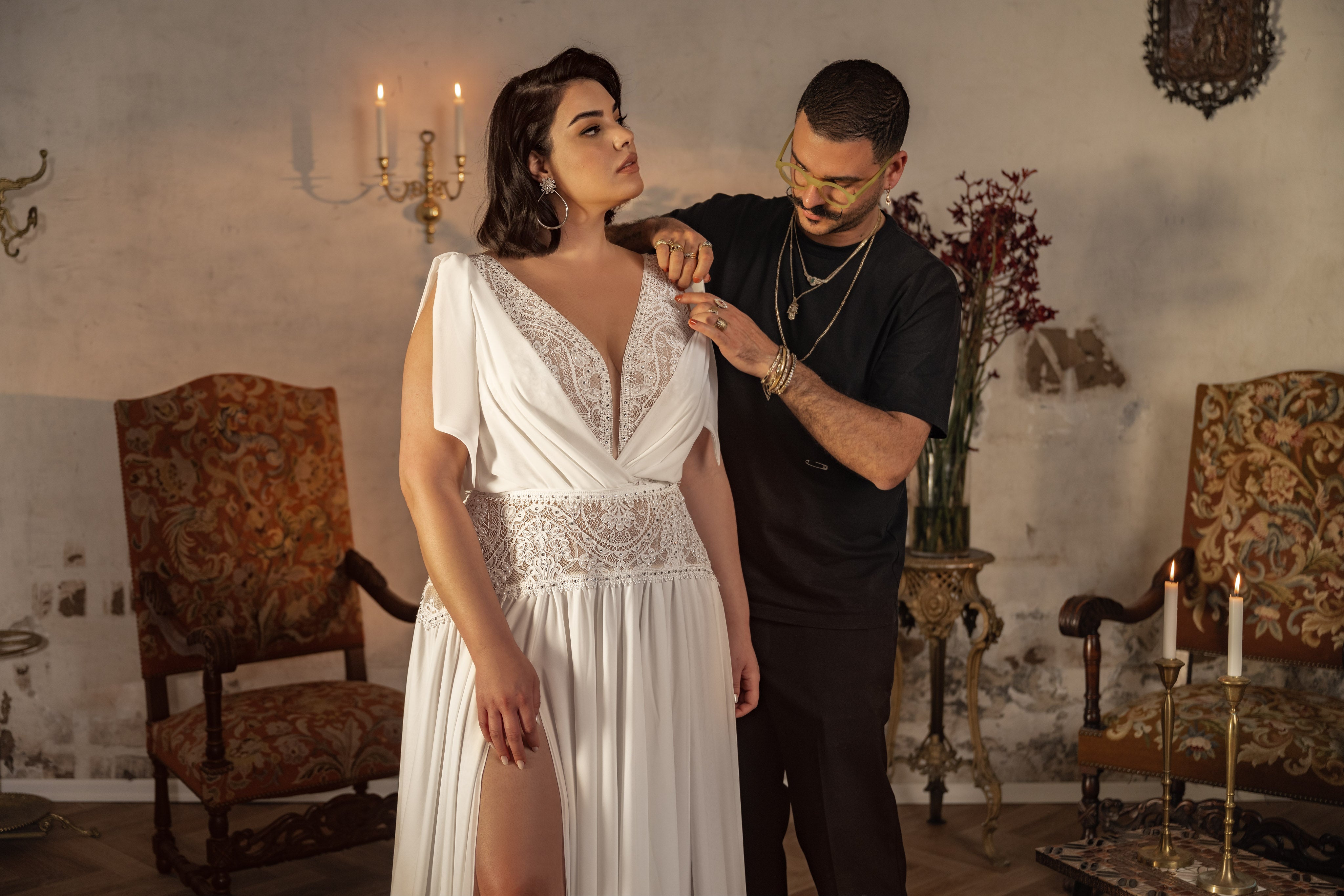 מעצב מתאים את מחשוף שמלת הכלה Kristen מקולקציית Bridal Plus 2023/24 בחדר וינטג' עם כיסאות מעוטרים, נרות ופרחים. האישה עומדת בשלווה בזמן שהמעצב מתרכז בהשלמת המראה שלה.