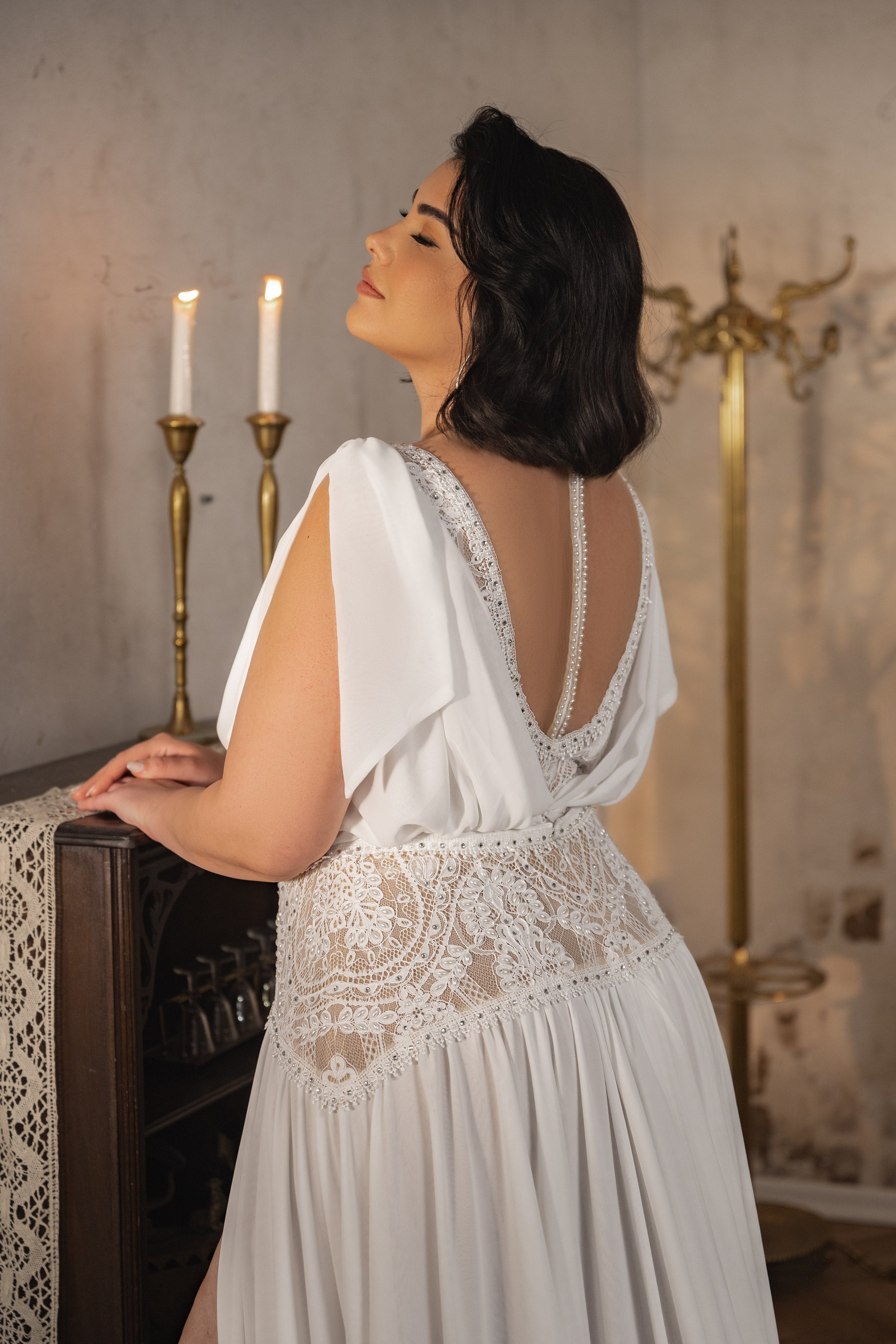 אישה מקולקציית Bridal Plus 2023/24 - שמלת קריסטן עומדת ליד ארון עץ מכוסה תחרה עם שני נרות פליז דולקים ומתלה מעילים, פונה לצד ומציגה את הגב הפתוח והאלגנטי של השמלה בעיניים עצומות.