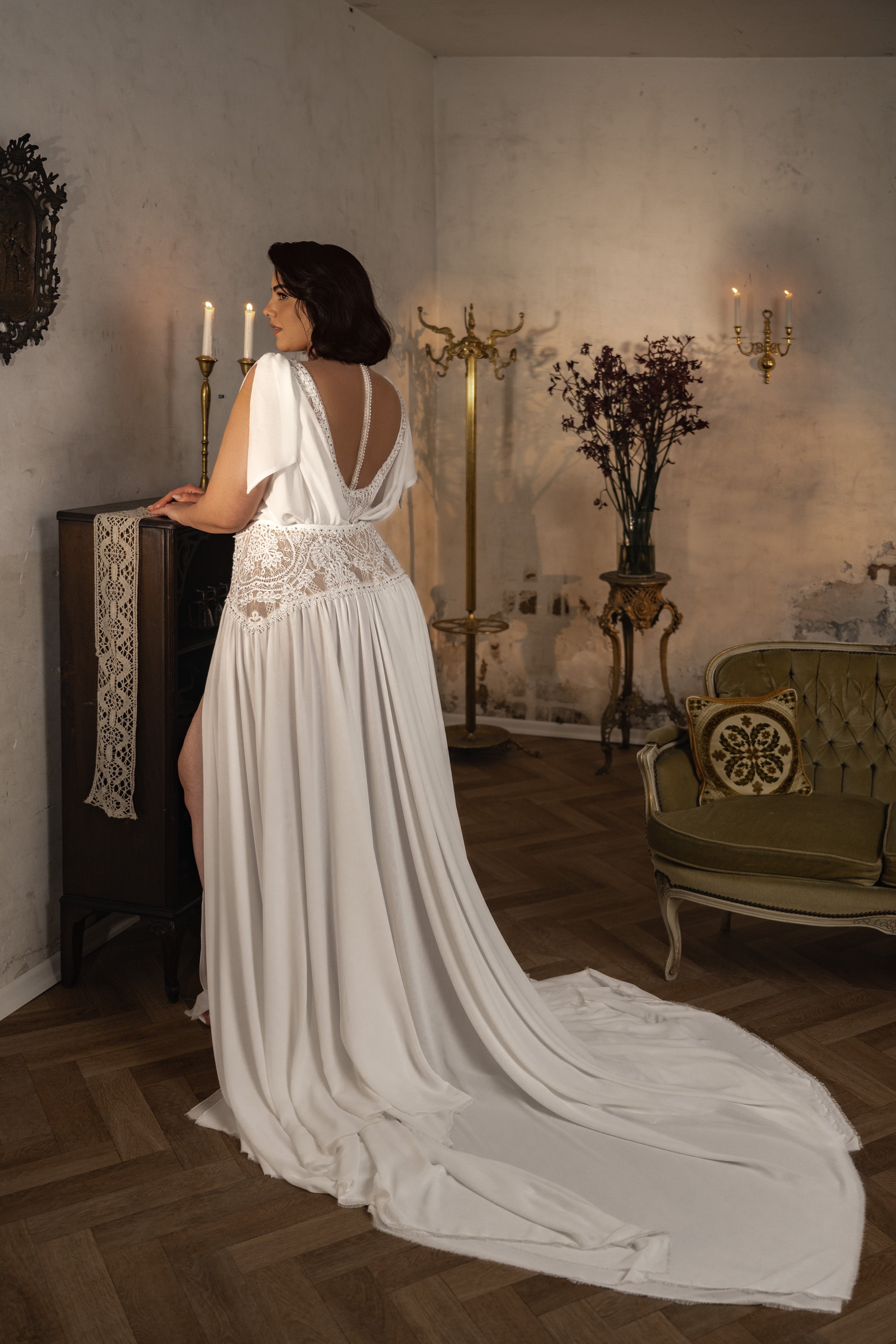 אישה לובשת שמלת כלה מקולקציית Bridal Plus 2023/24 - Kristen, עומדת ליד ארון בחדר בסגנון וינטג' עם נרות, נגיעות זהב, כיסא עתיק וסידור פרחים.