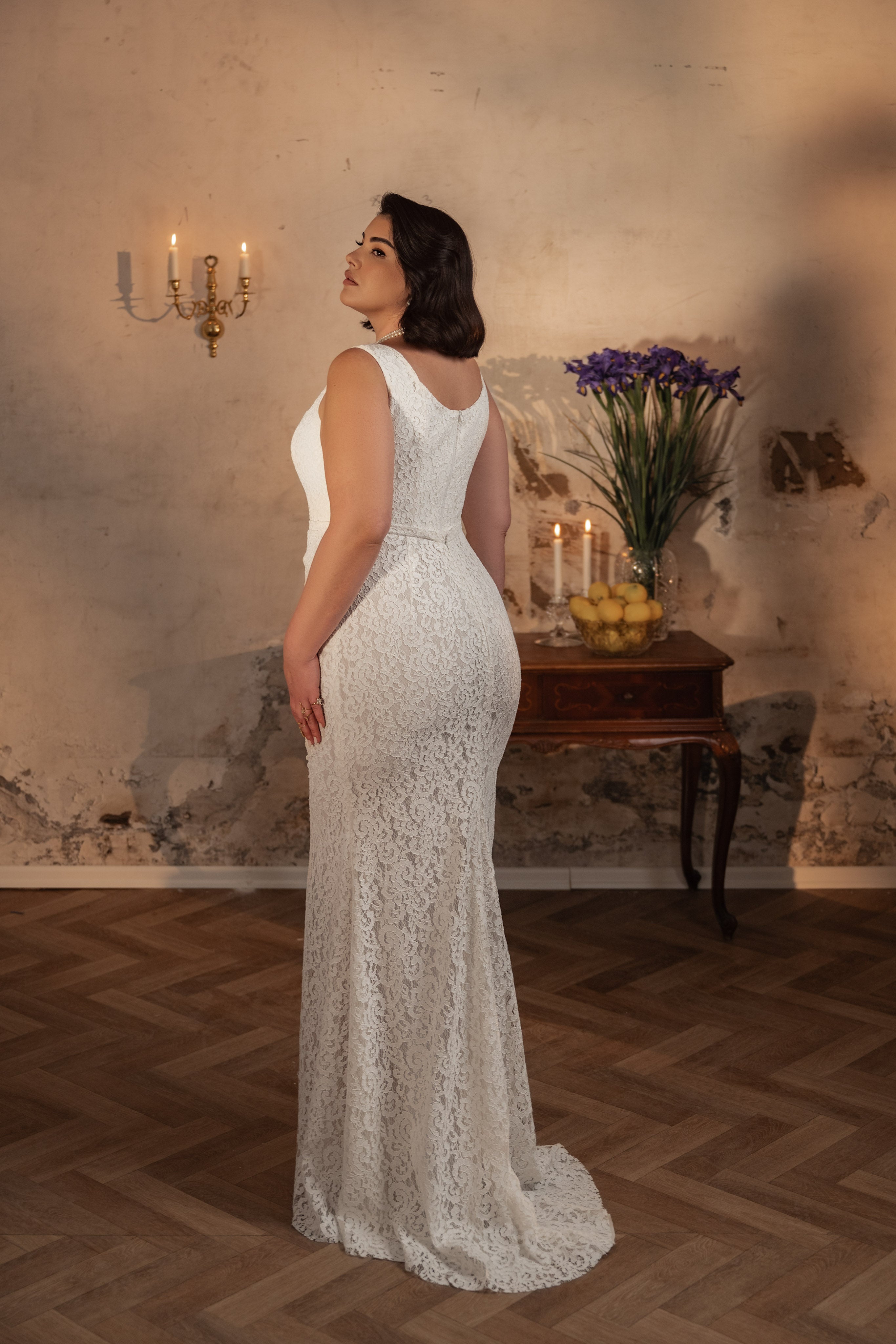 אישה לבושה בשמלת תחרה ללא שרוולים "Bridal Plus Collection 2023/24 - Irene" של Dror Kontento עומדת בתוך הבית ומביטה מעבר לכתפה. מאחוריה שולחן וינטג' עם נרות, לימונים, פרחים סגולים ופמוט קיר.