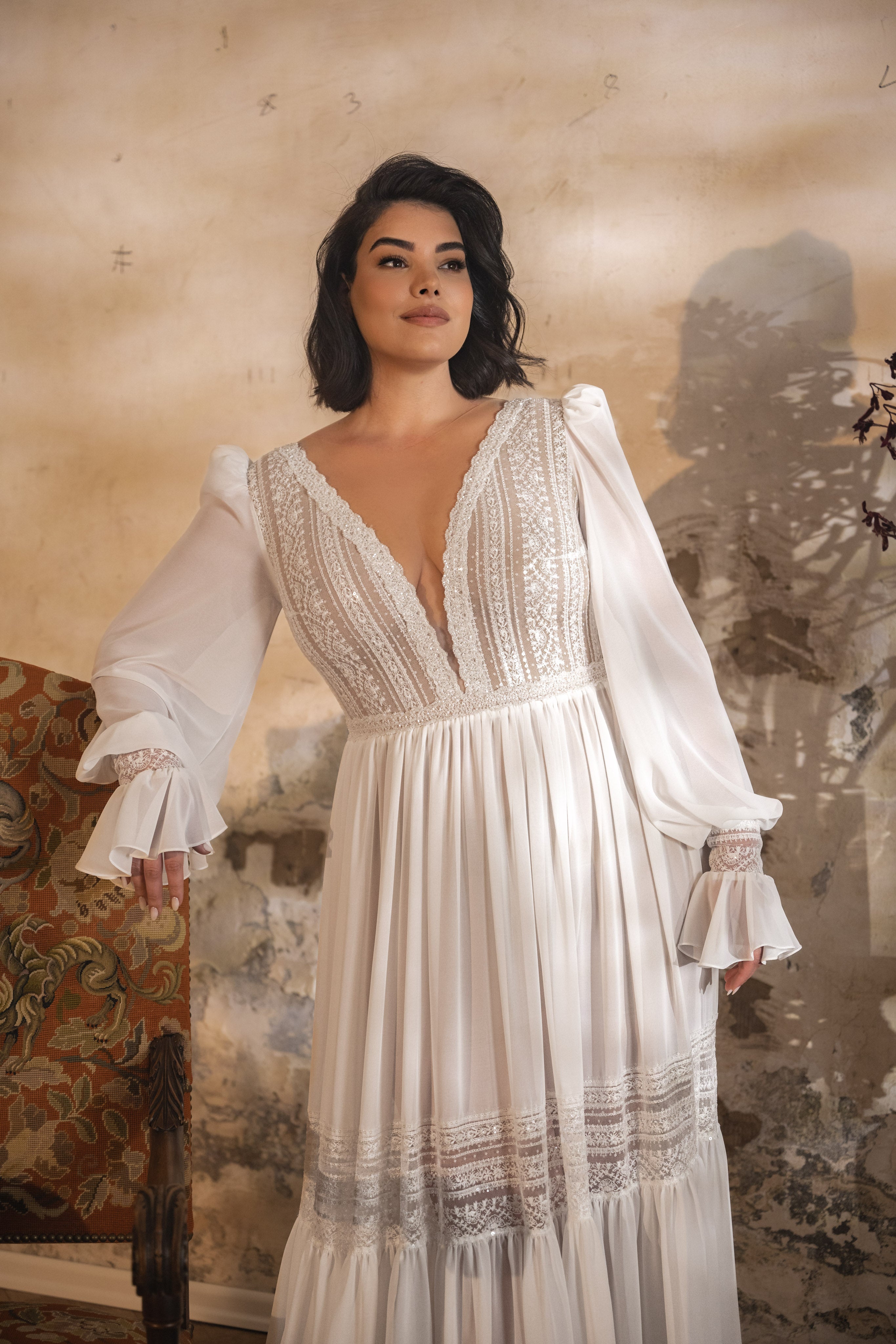 אישה עם שיער כהה לובשת שמלת כלה מקולקציית Bridal Plus 2023/24 - שמלת תחרה ושיפון Lilly-Rose, עומדת בתוך הבית ליד כיסא פרחוני וינטג' על רקע קיר כפרי ומחוספס.