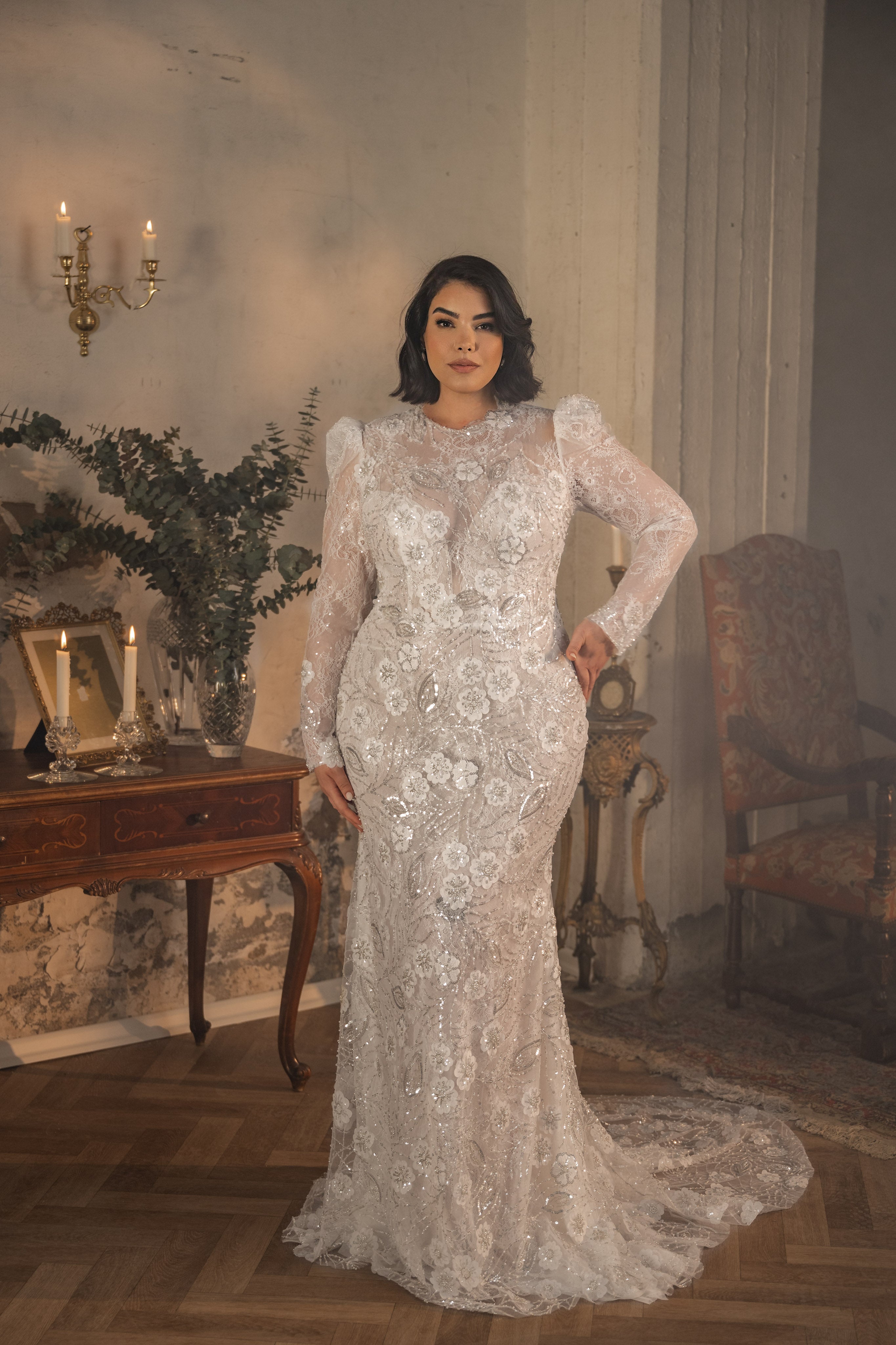 אישה מקולקציית Bridal Plus 2023/24 - שמלת כלה עם שרוולים ארוכים מתחרה של Shelby עומדת בתוך חדר אלגנטי ומואר באור רך, ליד שולחן עץ מעוטר בנרות וצמחייה.