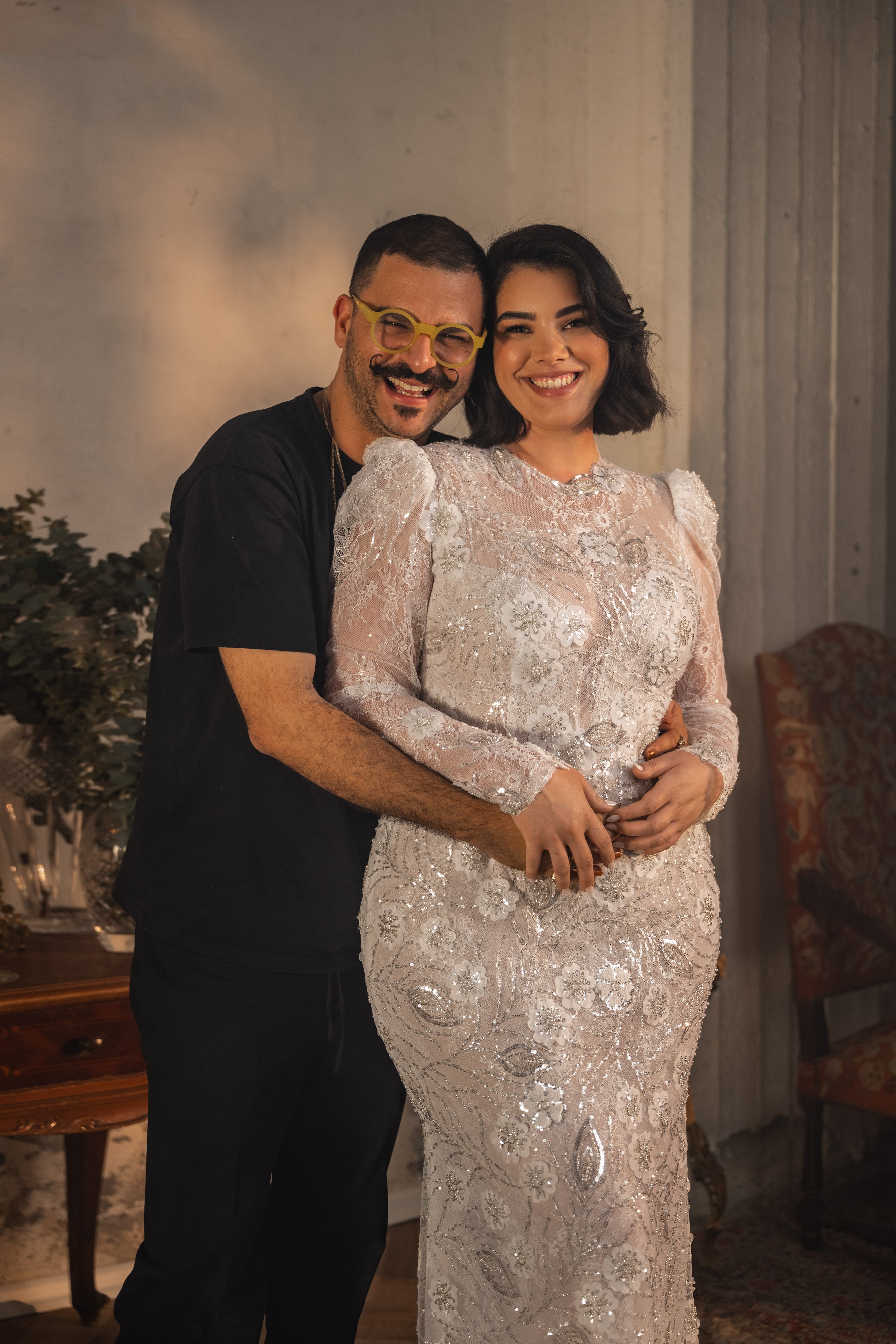 גבר לבוש שחור עם משקפיים צהובים מחבק אישה מחייכת הלובשת שמלת כלה מתחרת Bridal Plus Collection 2023/24 - Shelby. הם עומדים בתוך הבית ליד שולחן עץ עם צמחייה וכיסא מרופד.