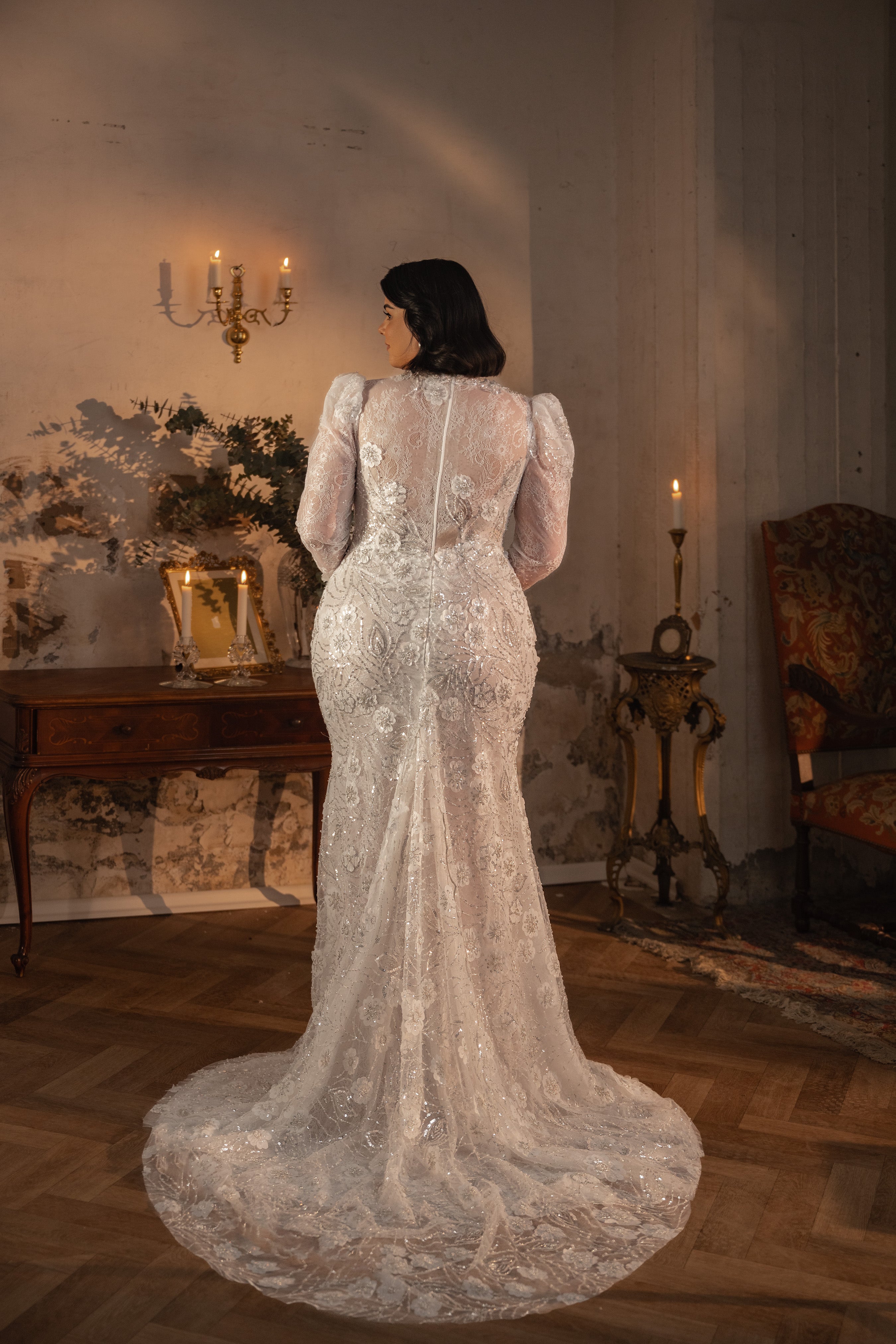 אישה מקולקציית Bridal Plus 2023/24 - שמלת כלה עם שרוולים ארוכים מתחרה של Shelby עומדת עם גבה למצלמה בחדר וינטג' המעוצב בנרות, שולחן עתיק ורהיטים מעוטרים.