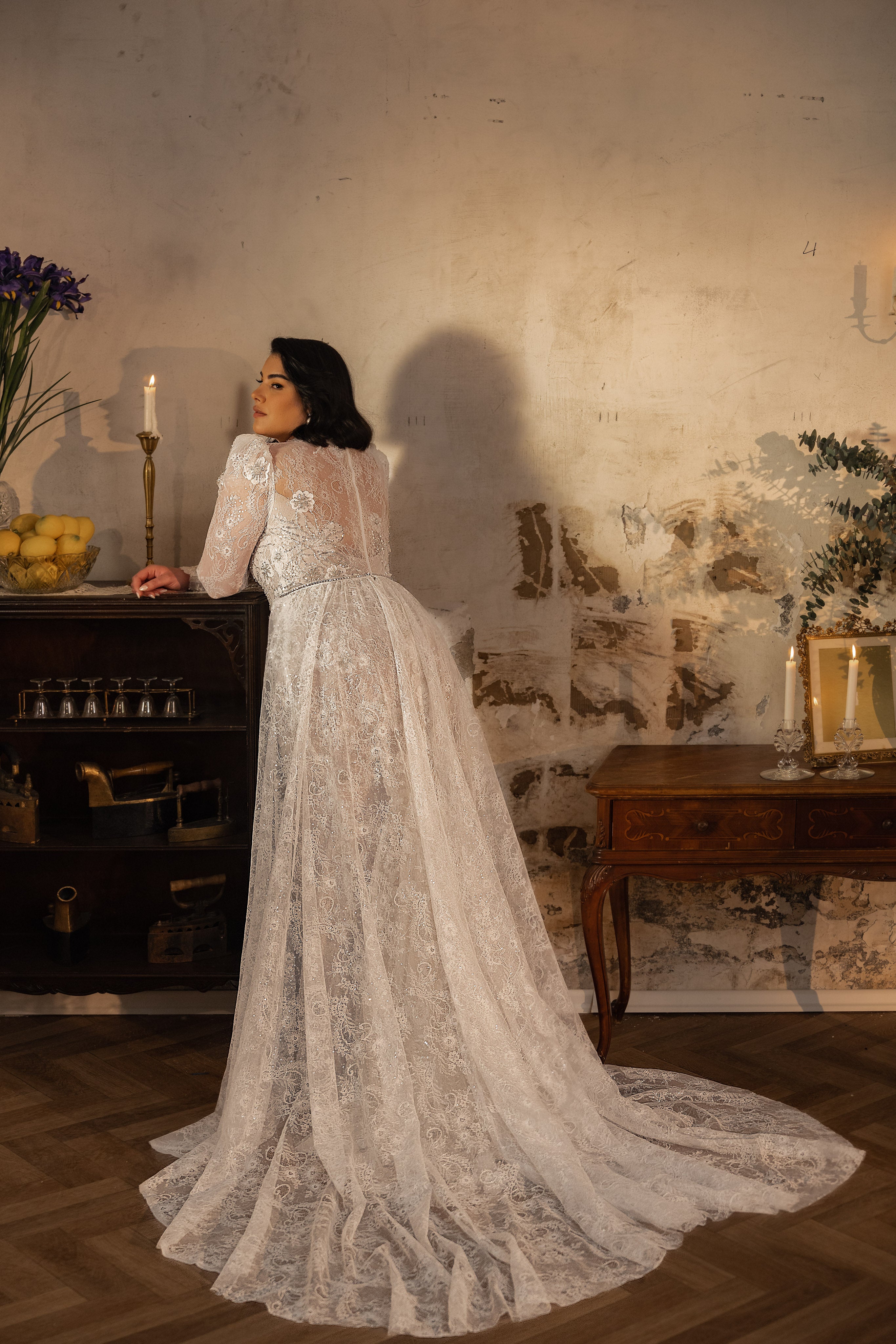 אישה בשמלת כלה מקולקציית Bridal Plus 2023/24 - Shelby נשענת על ארון וינטג' בחדר מואר באור רך עם קירות כפריים, פרחים ונרות, היוצרים אווירה רומנטית ואלגנטית.