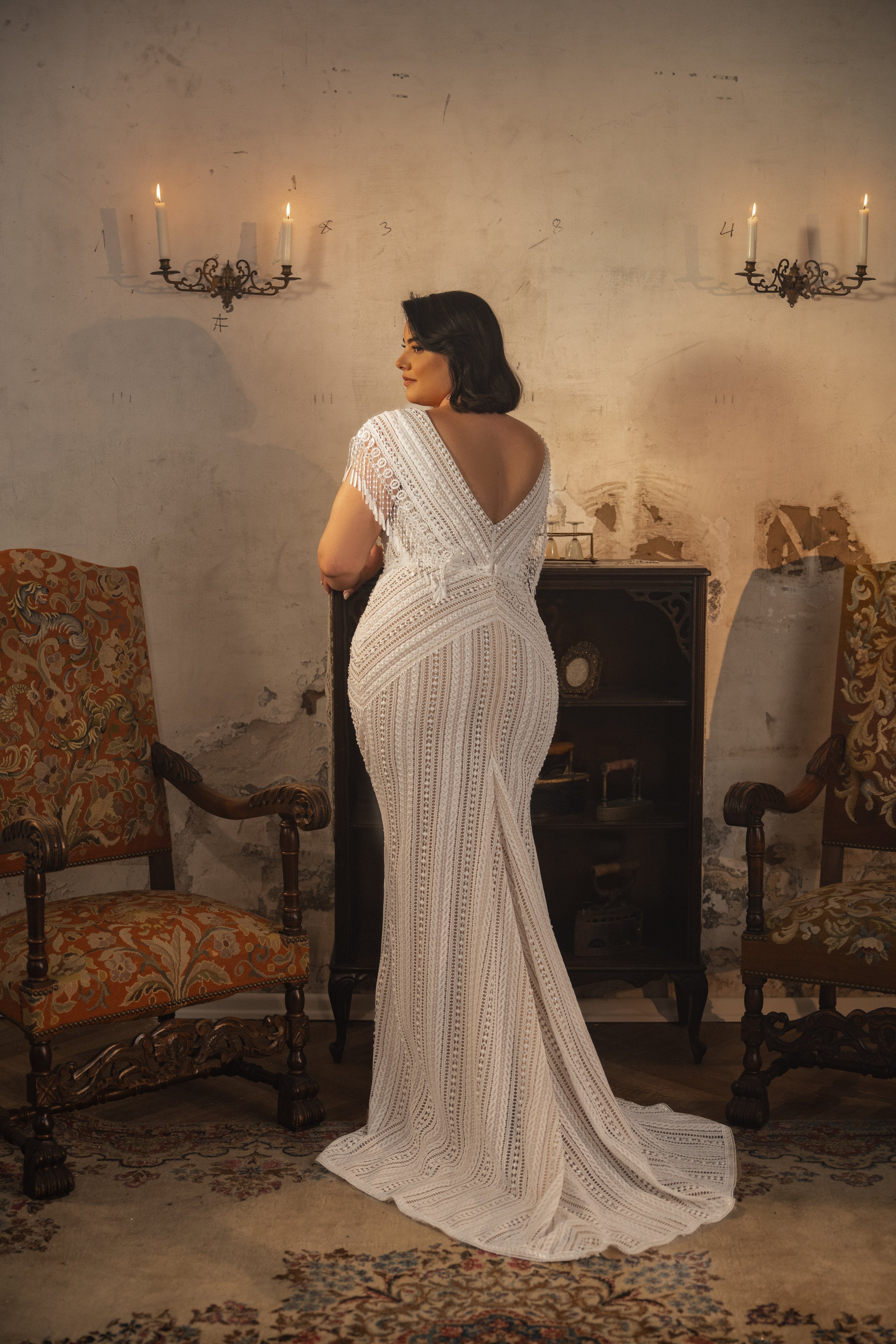 אישה מקולקציית Bridal Plus 2023/24 - שמלת Martina, עם פרטים מעוטרים ביד וחתך V עמוק בגב, עומדת עם גבה למצלמה בחדר וינטג' עם כיסאות מעוטרים, שטיח בדוגמה, קירות בלוים ופמוטים קיר מוארים.