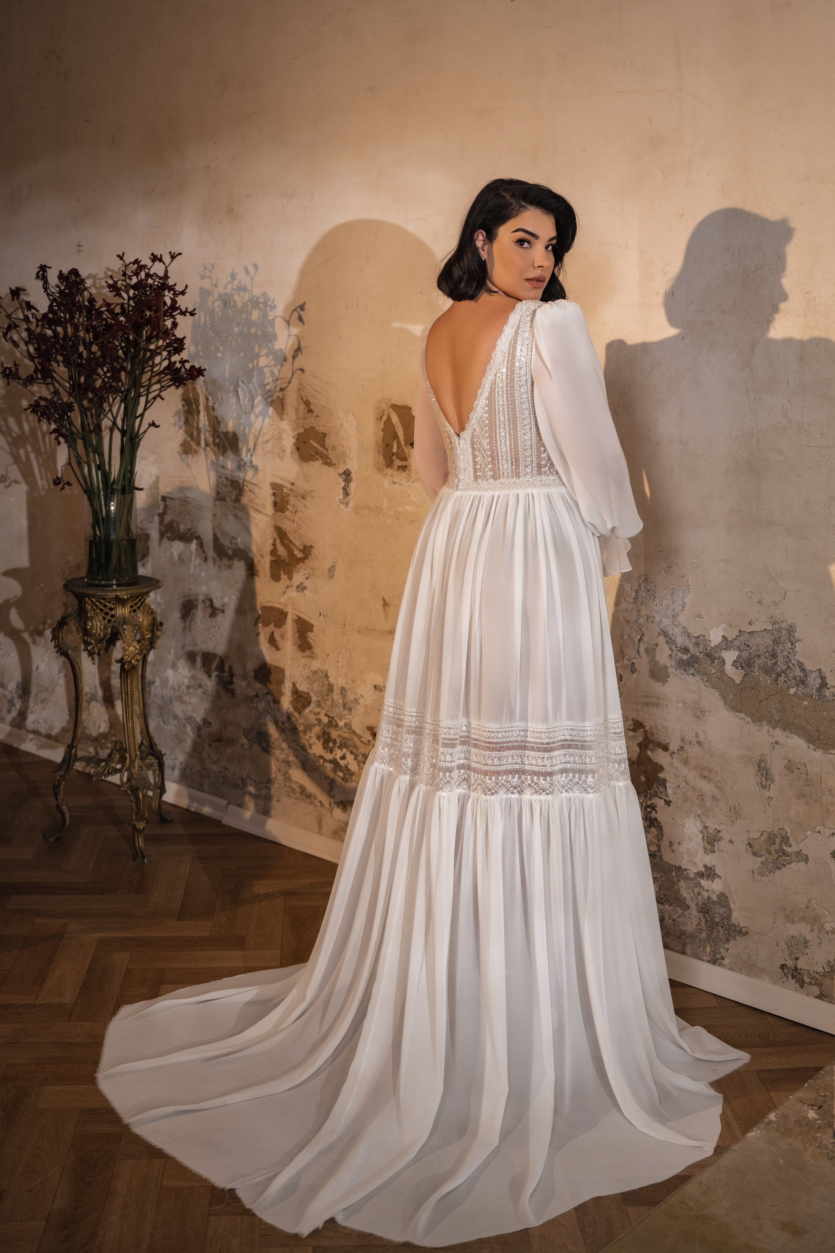 אישה לובשת את שמלת Lilly-Rose מקולקציית Bridal Plus 2023/24, עומדת בתוך הבית ליד קיר מרקם ושולחן מעוטר בפרחים אדומים כהים, כאשר האור מטיל צל אלגנטי מאחוריה.