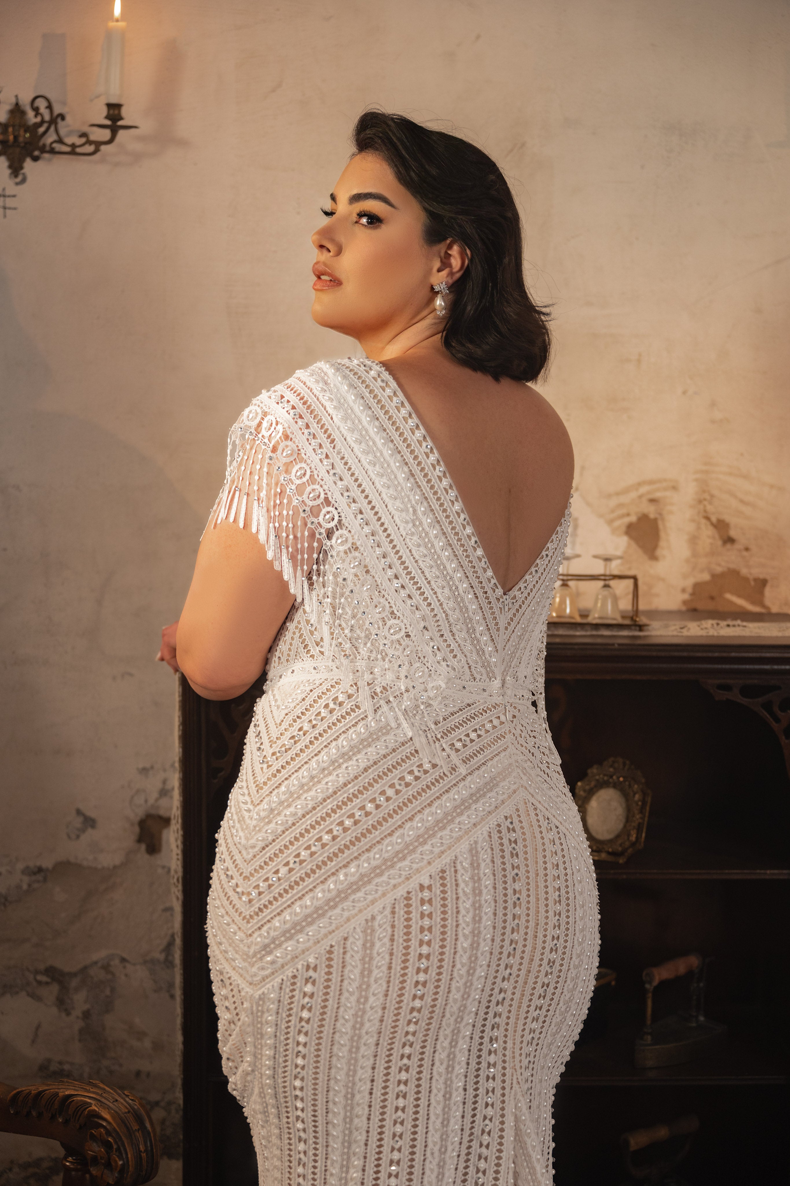 אישה עם שיער כהה מדגמנת את שמלת Martina מקולקציית Bridal Plus 2023/24, ומציגה את החרוזים האלגנטיים והשרוולים עם הפרינג'ים, בעודה עומדת בחדר וינטג' ומביטה מעבר לכתפה לעבר המצלמה.