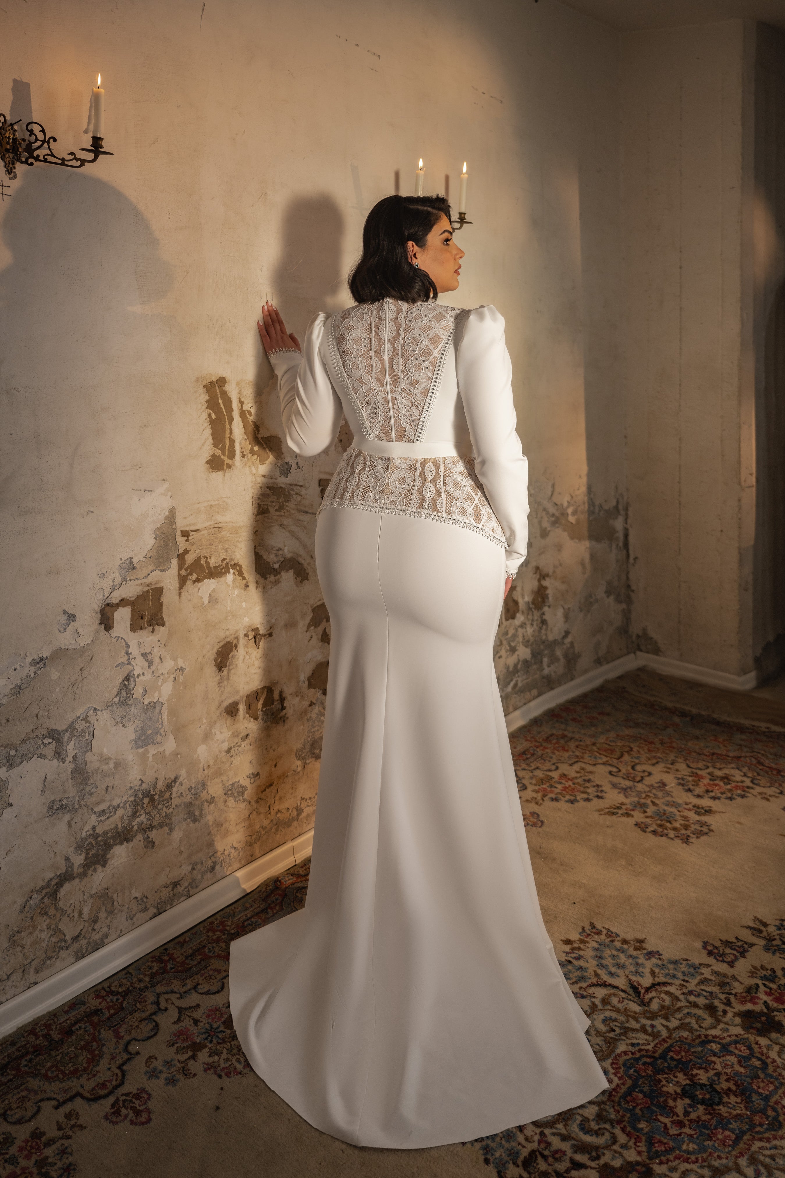 אישה בשמלת כלה ארוכה ומחמיאה מקולקציית Bridal Plus 2023/24–Vivian עומדת מול קיר מרקם ומיושן, וידה מונחת עליו. התפאורה כוללת שטיחים מעוצבים, פמוטי נרות ותאורה רכה וחמימה.