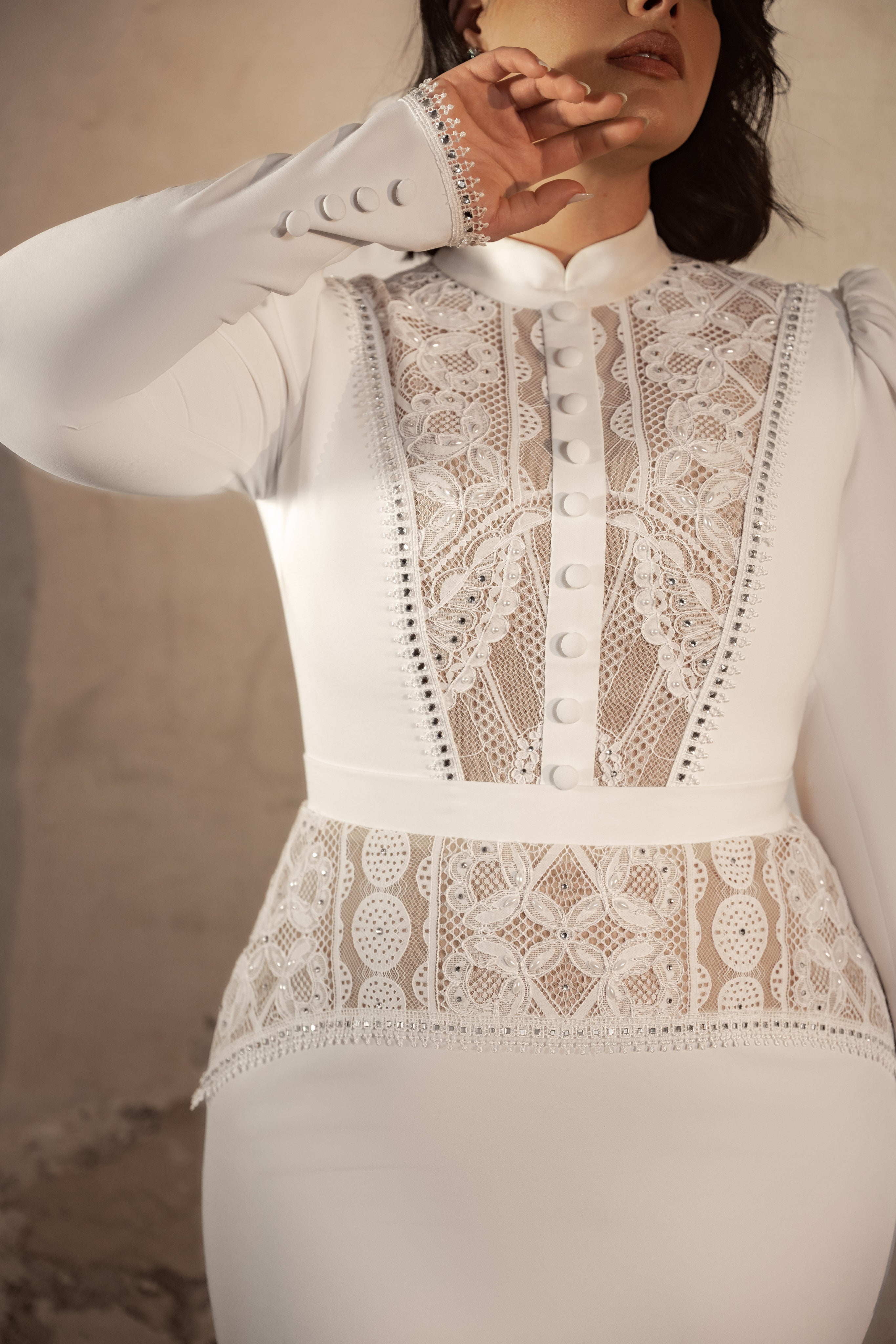 אישה לבושה בשמלת Vivian מקולקציית Bridal Plus 2023/24, עם פאנלים מחויטים מתחרה לבנה, כפתורים דקורטיביים ושרוולים ארוכים, מצטלמת כשכף ידה מכסה חלקית את פניה על רקע ניטרלי מואר באור רך.