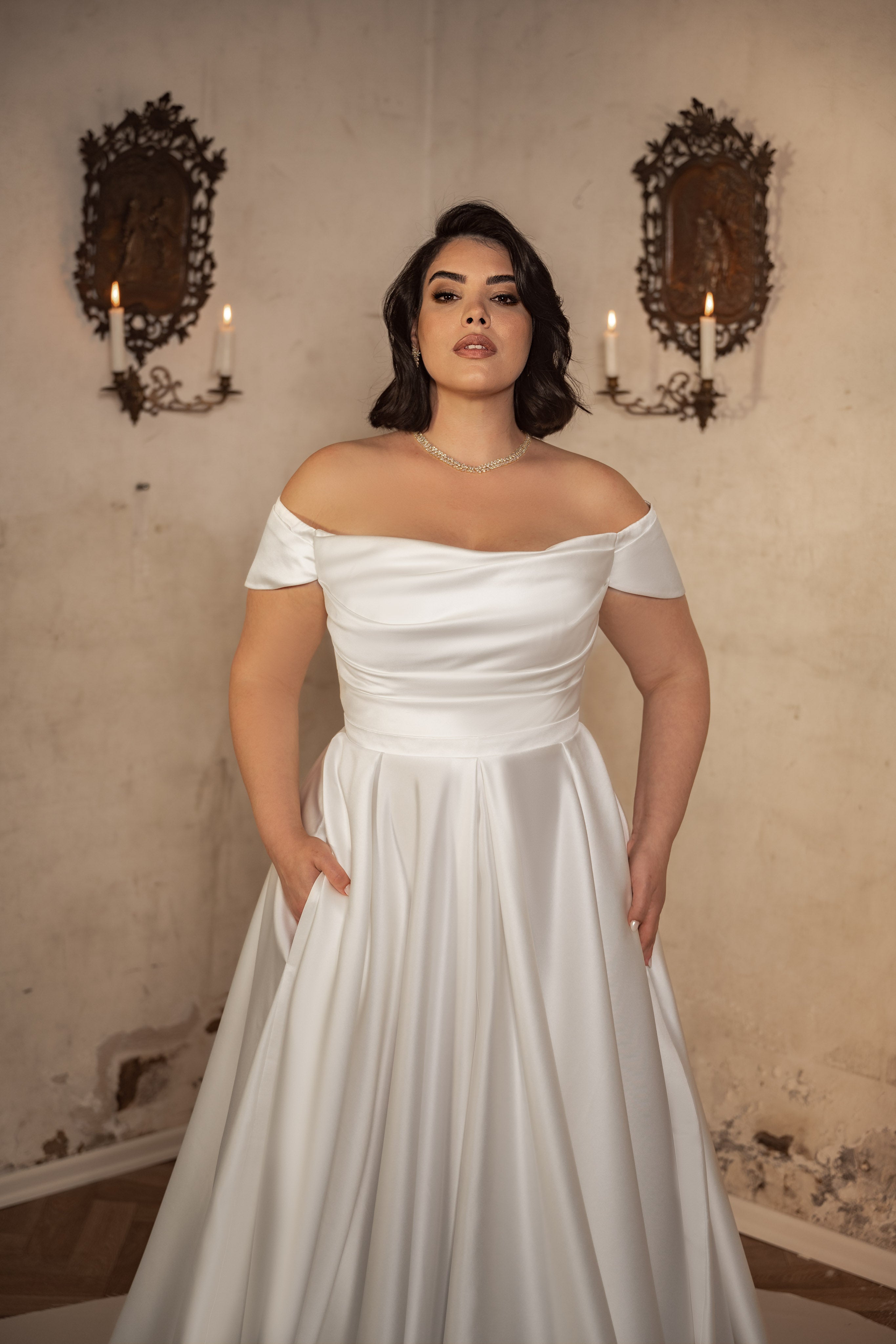 אישה מקולקציית Bridal Plus 2023/24 - שמלת כלה עם כתפיים חשופות מדגם Alicia עומדת בביטחון מול קיר בז' מרקם עם פמוטי נרות, ומדגישה את האלגנטיות של שמלות כלה מבד סאטן קרפ במידות גדולות.