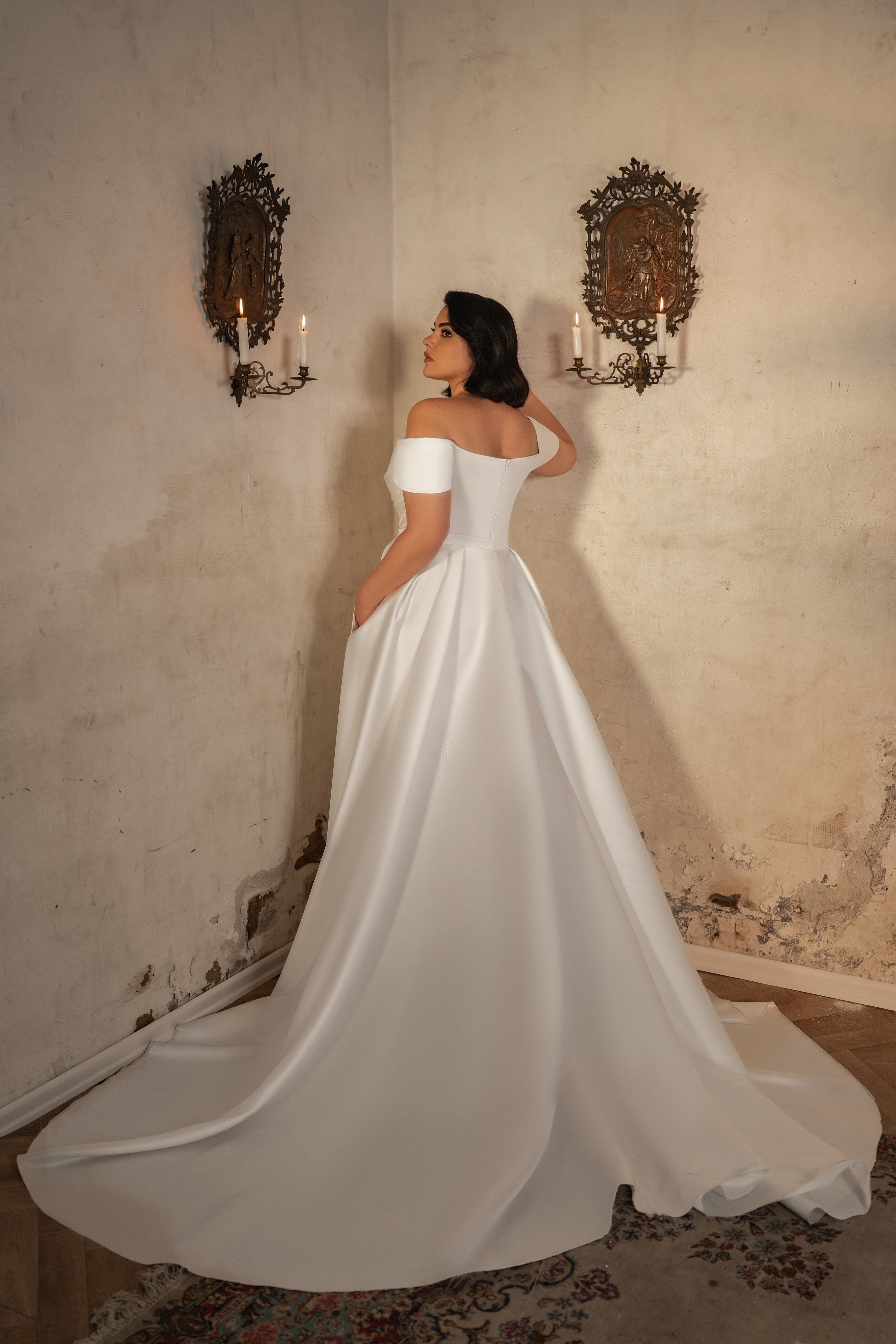 אישה לובשת שמלת כלה מהקולקציה Bridal Plus 2023/24 - Alicia עם כתפיים חשופות ושובל ארוך, עומדת בחדר וינטג' עם פמוטי נרות מעוטרים וקירות מרקמים במראה מיושן.