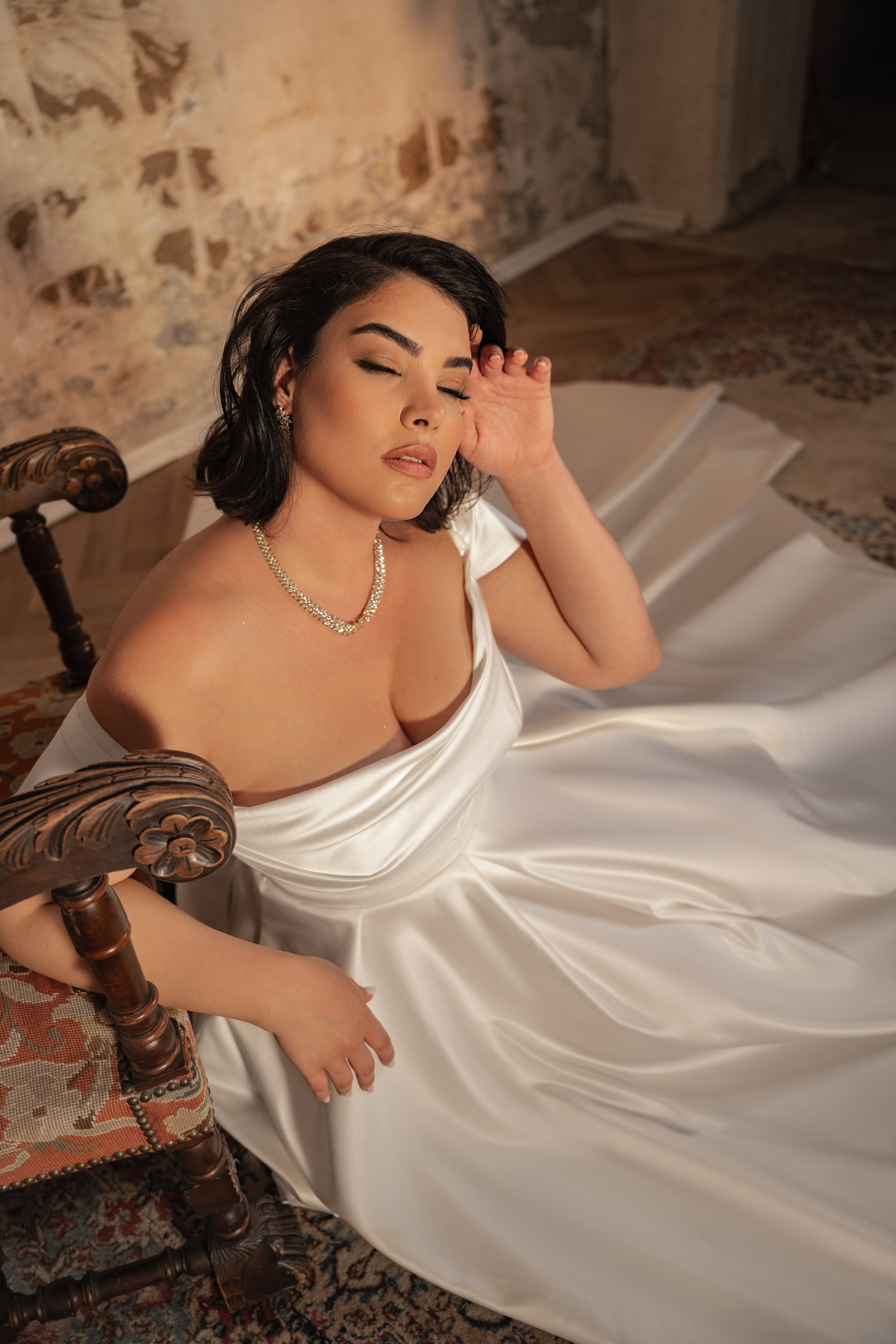 אישה מקולקציית Bridal Plus 2023/24 - שמלת כלה עם כתפיים חשופות של Alicia יושבת על הרצפה ליד כיסא עץ, עיניה עצומות וידה ליד פניה, כאשר אור רך מסנן דרך חדר כפרי עם קירות לבנים חשופים ושטיחים מעוצבים.