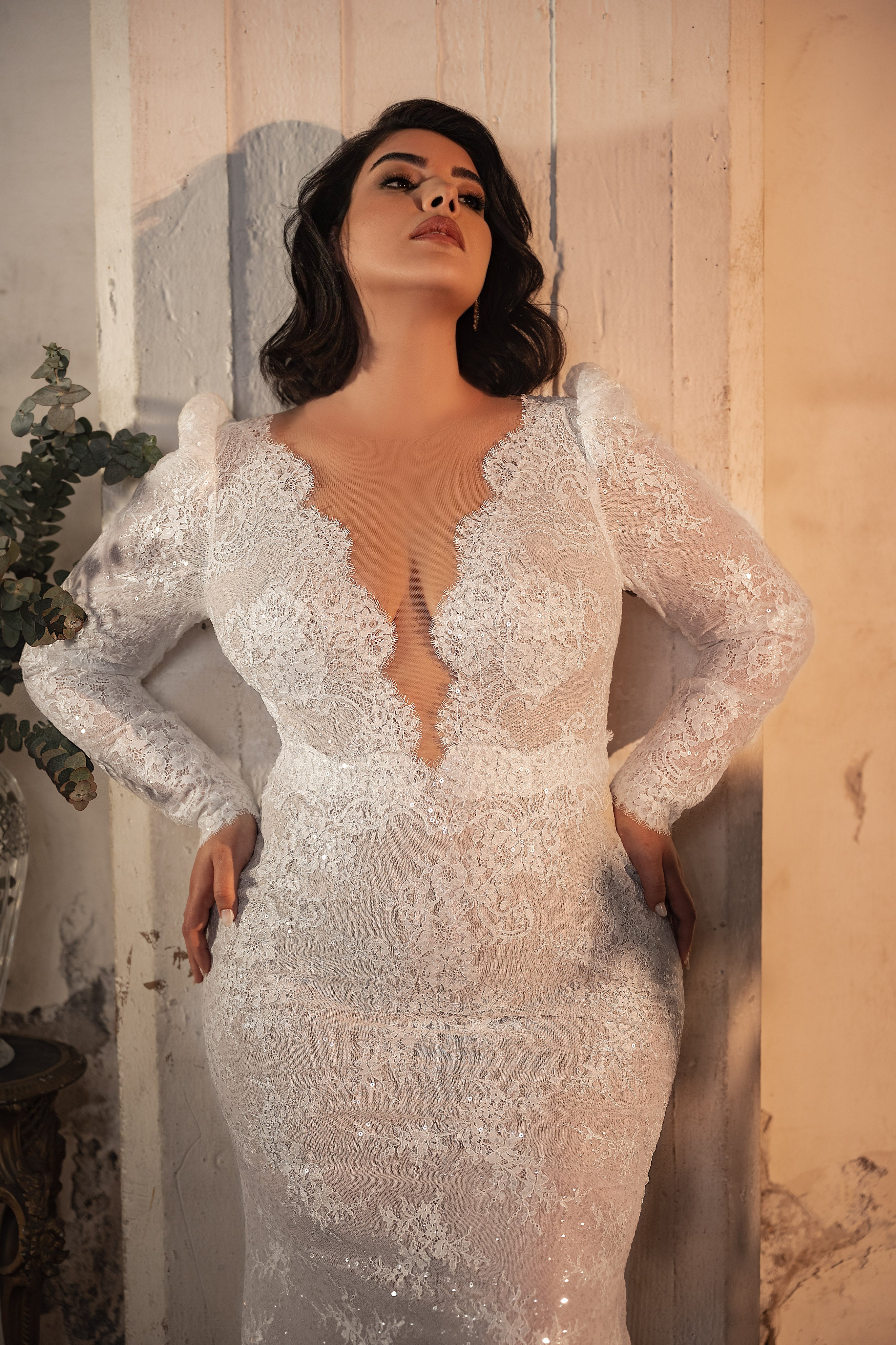 אישה לבושה בשמלת כלה מהקולקציה Bridal Plus Collection 2023/24 - Hope, שמלת תחרה לבנה עם שרוולים ארוכים, עומדת מול קיר בעל מרקם בהיר, מביטה למעלה וידיה על מותניה — דוגמה אלגנטית לשמלות כלה במידות גדולות.