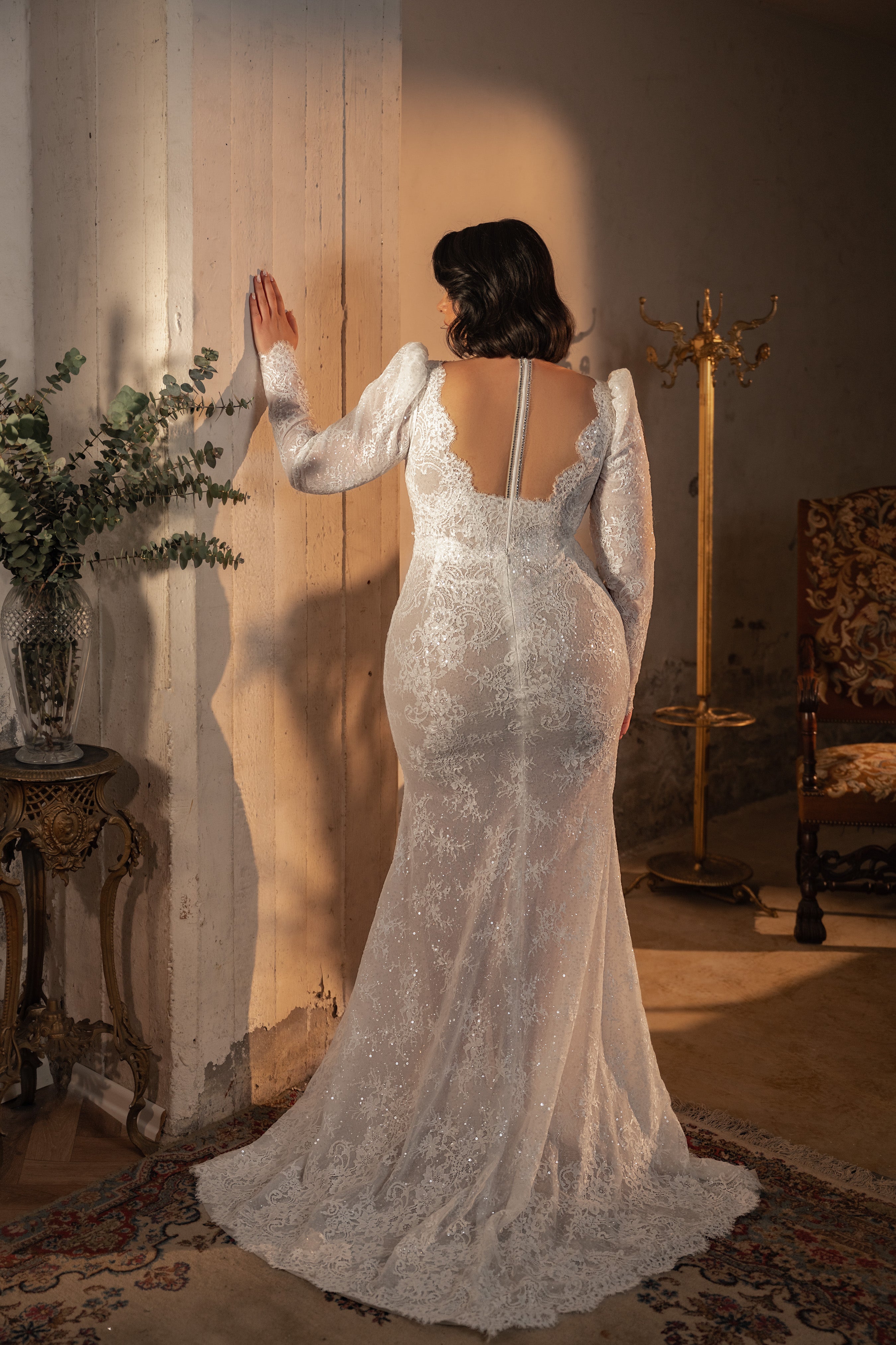 אישה בשמלת תחרה עם שרוולים ארוכים מקולקציית Bridal Plus 2023/24 - Hope עומדת עם גבה למצלמה, נוגעת בקיר בחדר בסגנון וינטג' מואר באור חם, עם ריהוט מעוטר ושטיח דקורטיבי.