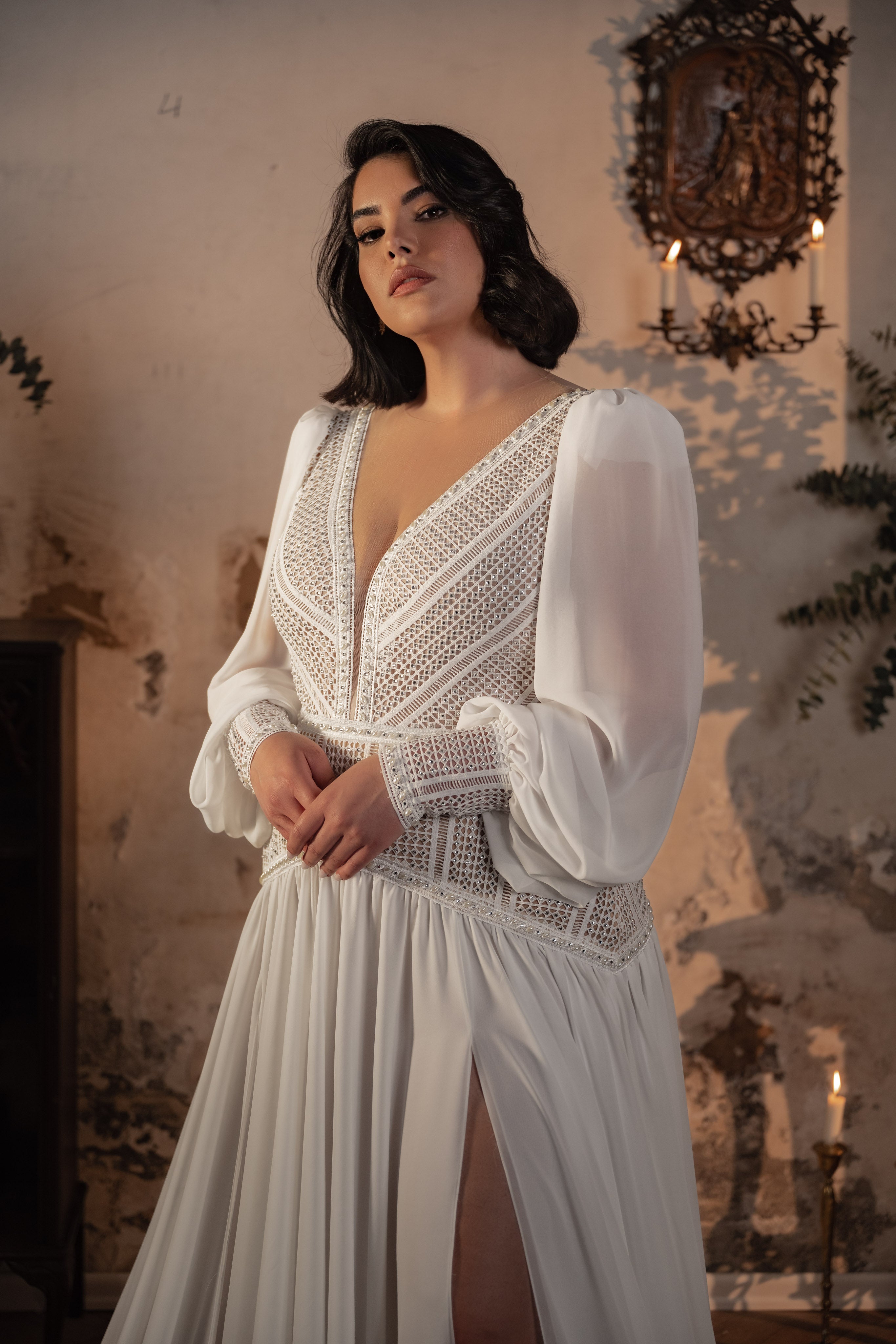 אישה מקולקציית Bridal Plus 2023/24 - שמלת Georgina, עם חרוזים, פרטים מתחרה ושרוולים שקופים, עומדת בתוך הבית על רקע קיר מרקם כפרי עם נרות ועלים ברקע.