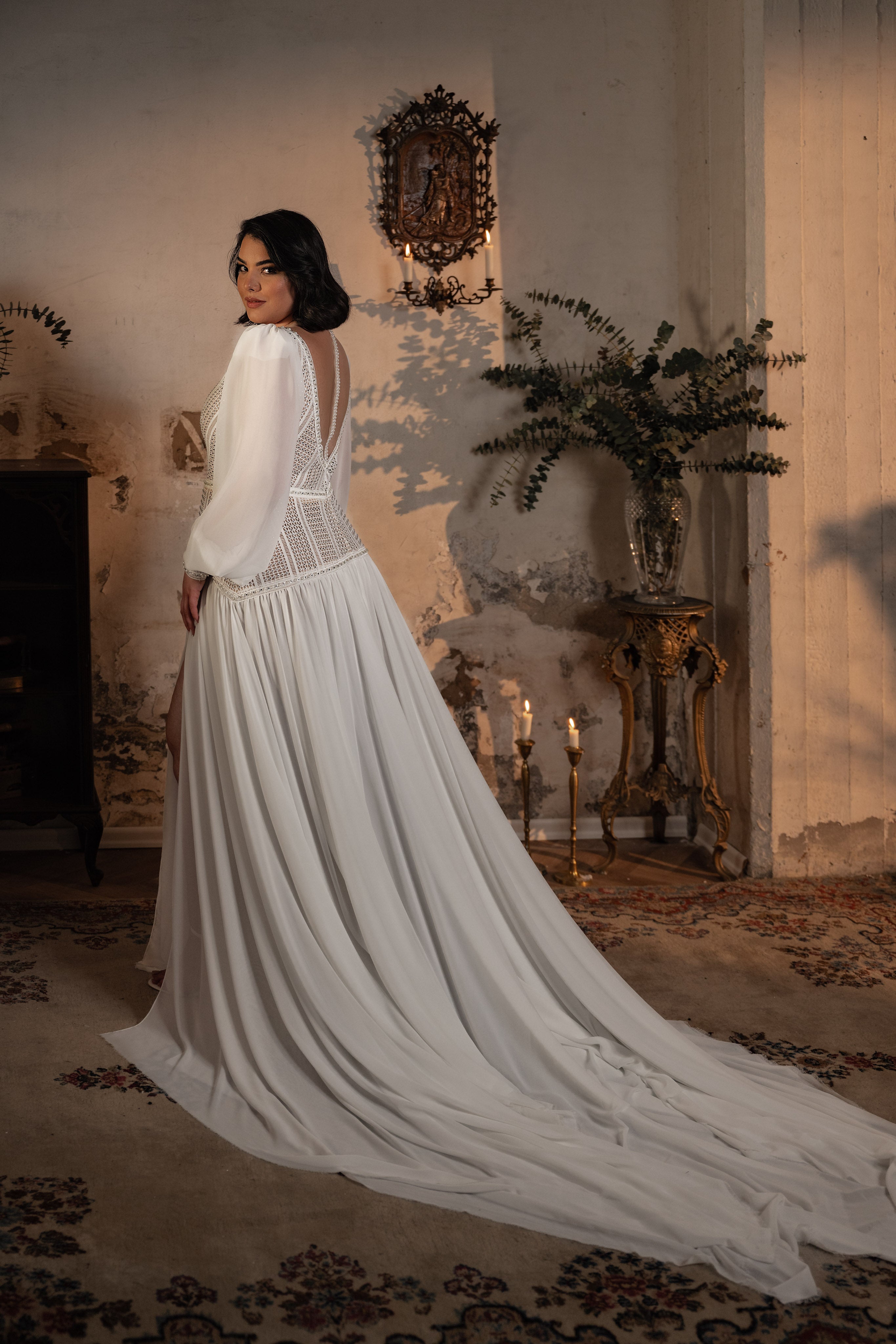 אישה לבושה בשמלת כלה מהקולקציה Bridal Plus Collection 2023/24 Georgina עומדת בחדר וינטג' עם קירות מרקמים, תפאורה עתיקה, נרות וענפים ירוקים, ומביטה מעבר לכתפה לעבר המצלמה.
