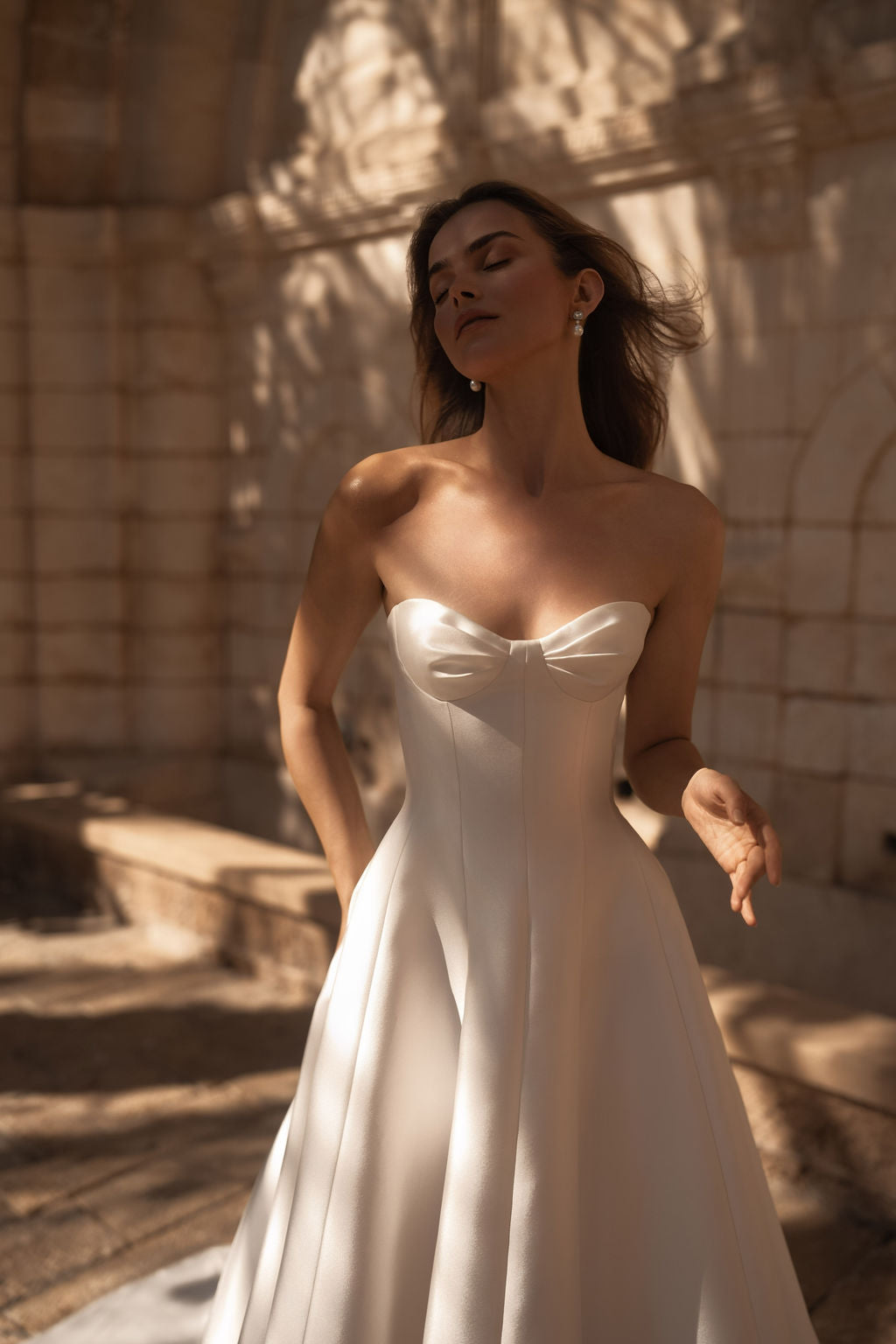 אישה לבושה בשמלת כלה NADYA W מקולקציית שמלות כלה – MID 2026 עומדת בחוץ, עיניה עצומות, בעוד אור השמש והצללים יוצרים דפוסים עדינים על שמלת הסאטן בצבע שנהב שלה, בעלת מחוך סטרפלס, ועל קיר האבן שמאחוריה. עיצוב מרהיב זה מספק הצצה יפהפייה לאלגנטיות הנצחית של שמלות כלה 2026.