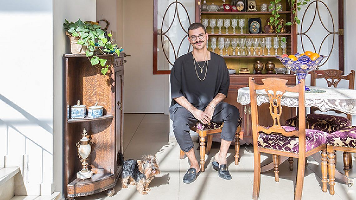 גבר עם משקפיים ושפם יושב על כיסא עץ בחדר אוכל מואר, עם כלב קטן לצידו. בחדר יש רהיטים וינטג', צמחים וויטרינה עם כלי זכוכית.