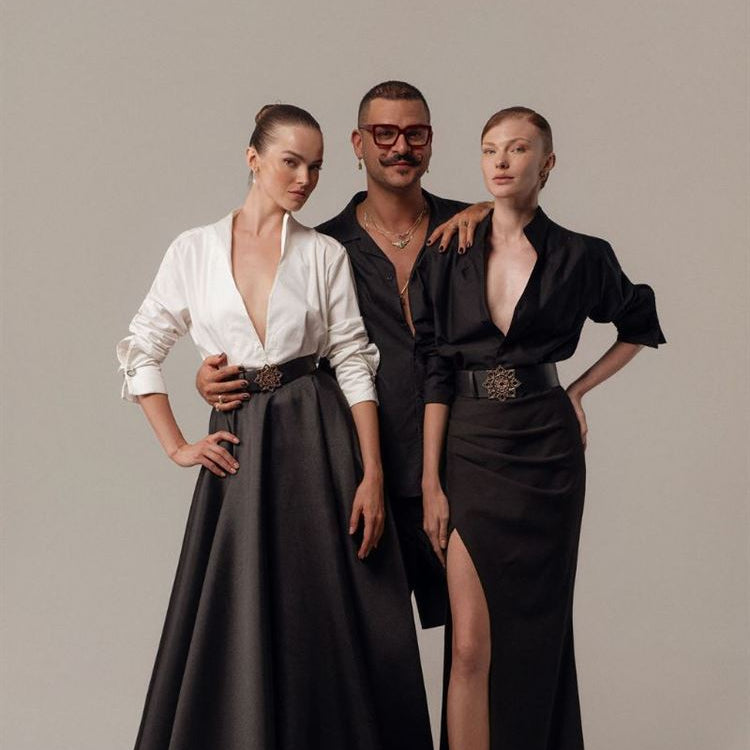 שלושה אנשים מצטלמים יחד על רקע אחיד. שתי נשים בחצאיות שחורות ארוכות וחולצות עם צווארון פתוח עומדות משני צדי גבר לבוש בבגדים כהים ומשקפיים. לכולם הבעת פנים ניטרלית ותלבושות מסוגננות.