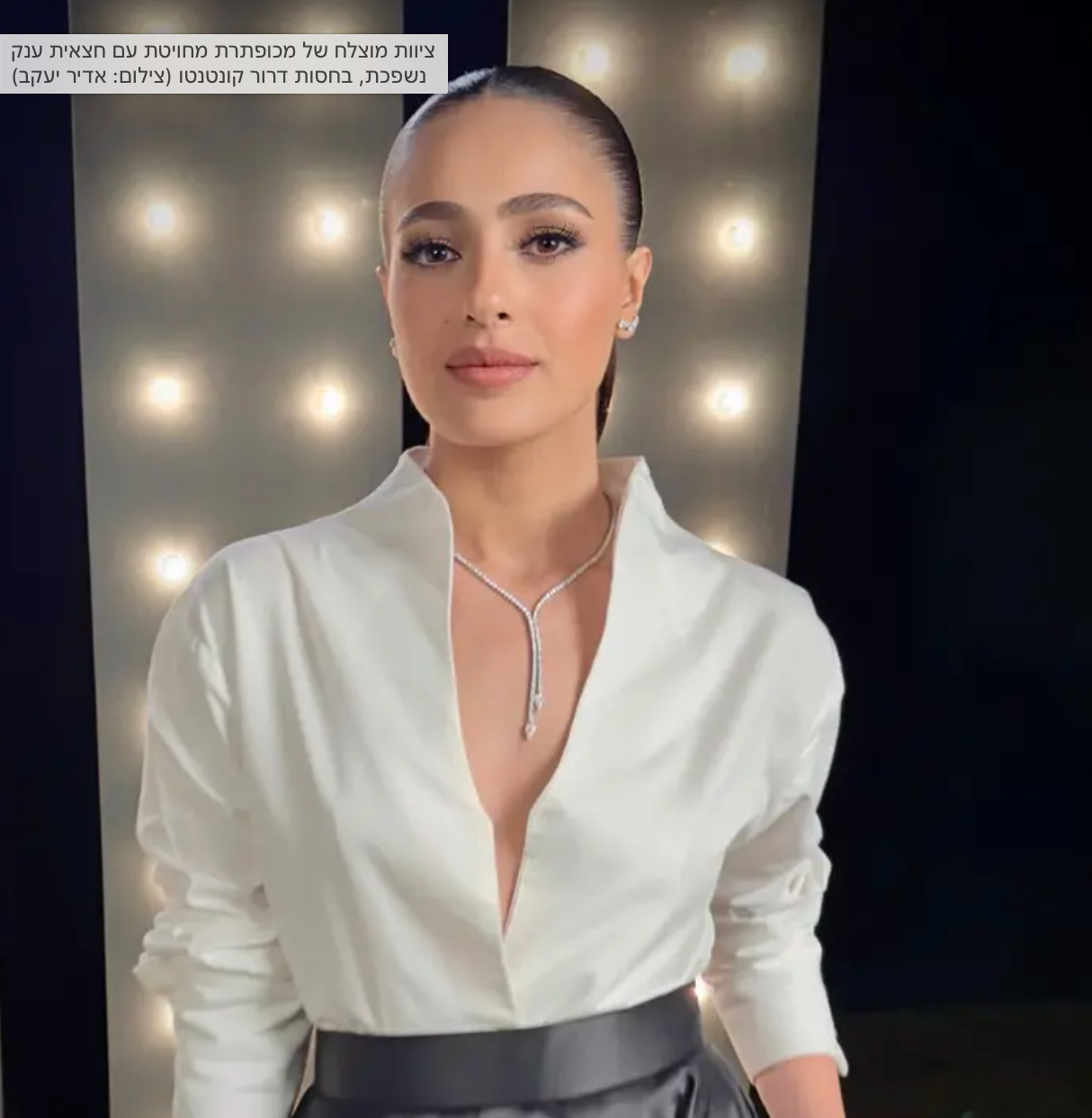 אישה עם שיער חלק, לבושה בחולצה לבנה וחצאית שחורה, עומדת מול רקע עם אורות בוהקים. היא עונדת שרשרת טיפה ומאופרת באיפור עדין, ומביטה בביטחון למצלמה.