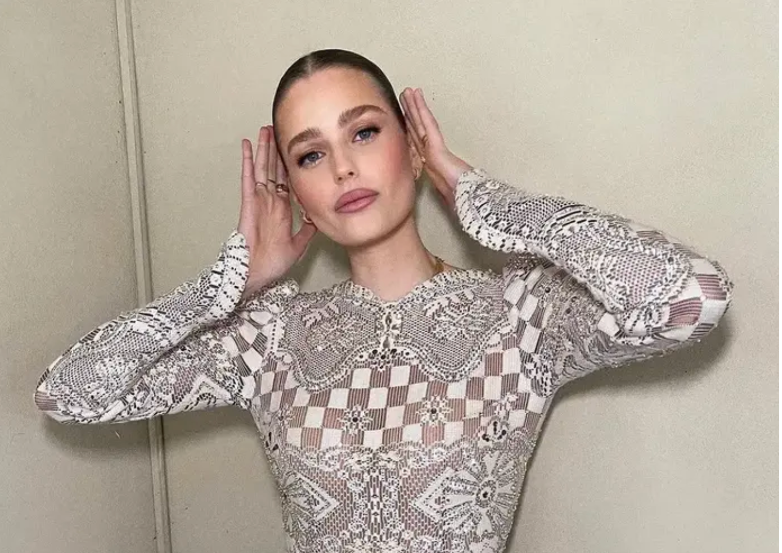 אישה עם שיער משוח לאחור מצטלמת עם ידיה ליד ראשה, לבושה בשמלה ארוכת שרוולים עם דוגמאות מורכבות, עומדת על רקע קיר בהיר וחלק.