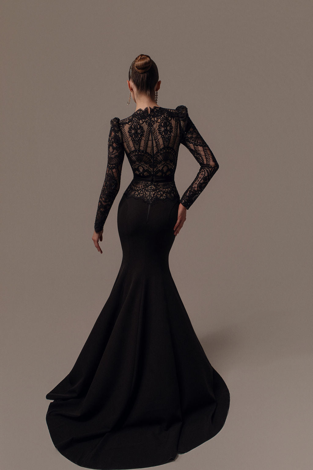 אישה עם שיער אסוף בצמה מצטלמת עם גבה למצלמה, לבושה בשמלת Leya מקולקציית ערב 2026 — שמלה שחורה אלגנטית עם תחרה וחצאית דרמטית בסגנון בת ים, על רקע חלק.
