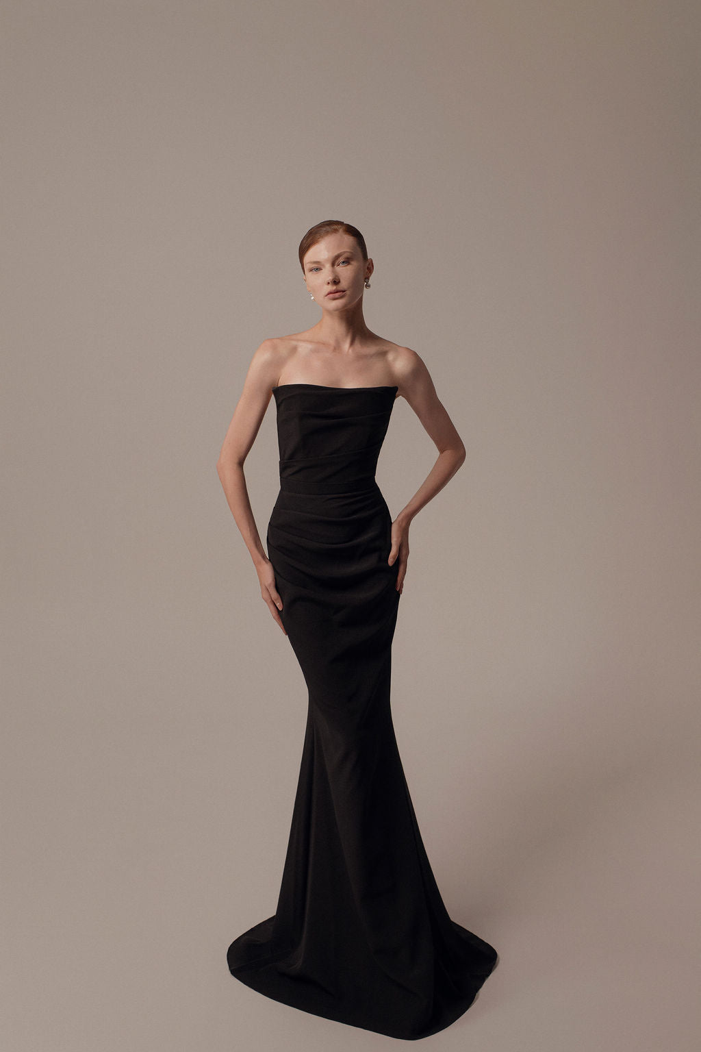 אישה מצטלמת על רקע ניטרלי בשמלת Nara מקולקציית הערב 2026 של דרור קונטנטו — שמלה שחורה ללא כתפיות, באורך הרצפה, המחבקת את גופה ומתרחבת כלפי מטה, עם שיער אסוף לאחור וידיים על המותניים.