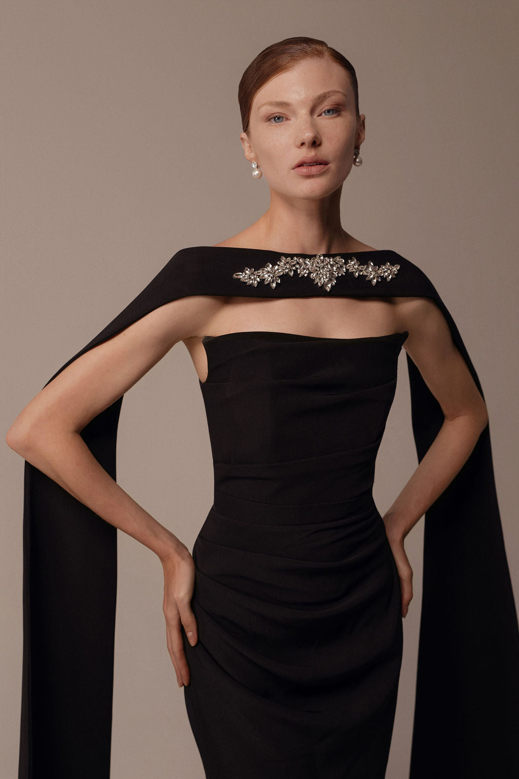 אישה עומדת בביטחון בשמלת Nara מקולקציית Evening 2026, שמלת ערב שחורה ואלגנטית ללא כתפיות עם גלימה דרמטית ומחשוף מעוטר בנצנצים, המשקפת את האלגנטיות המעודנת של דרור קונטנטו.