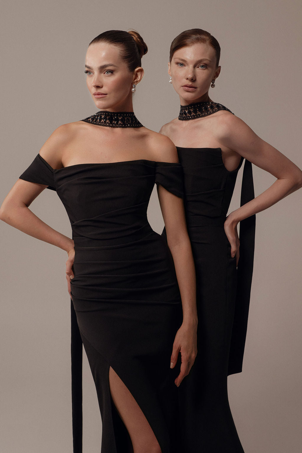 שתי נשים מצטלמות בביטחון בשמלות "רונה" אלגנטיות מקולקציית הערב 2026 של דרור קונטנטו — שמלות שחורות עם כתפיים חשופות, חתכים גבוהים ושרשראות מעוטרות — על רקע ניטרלי.