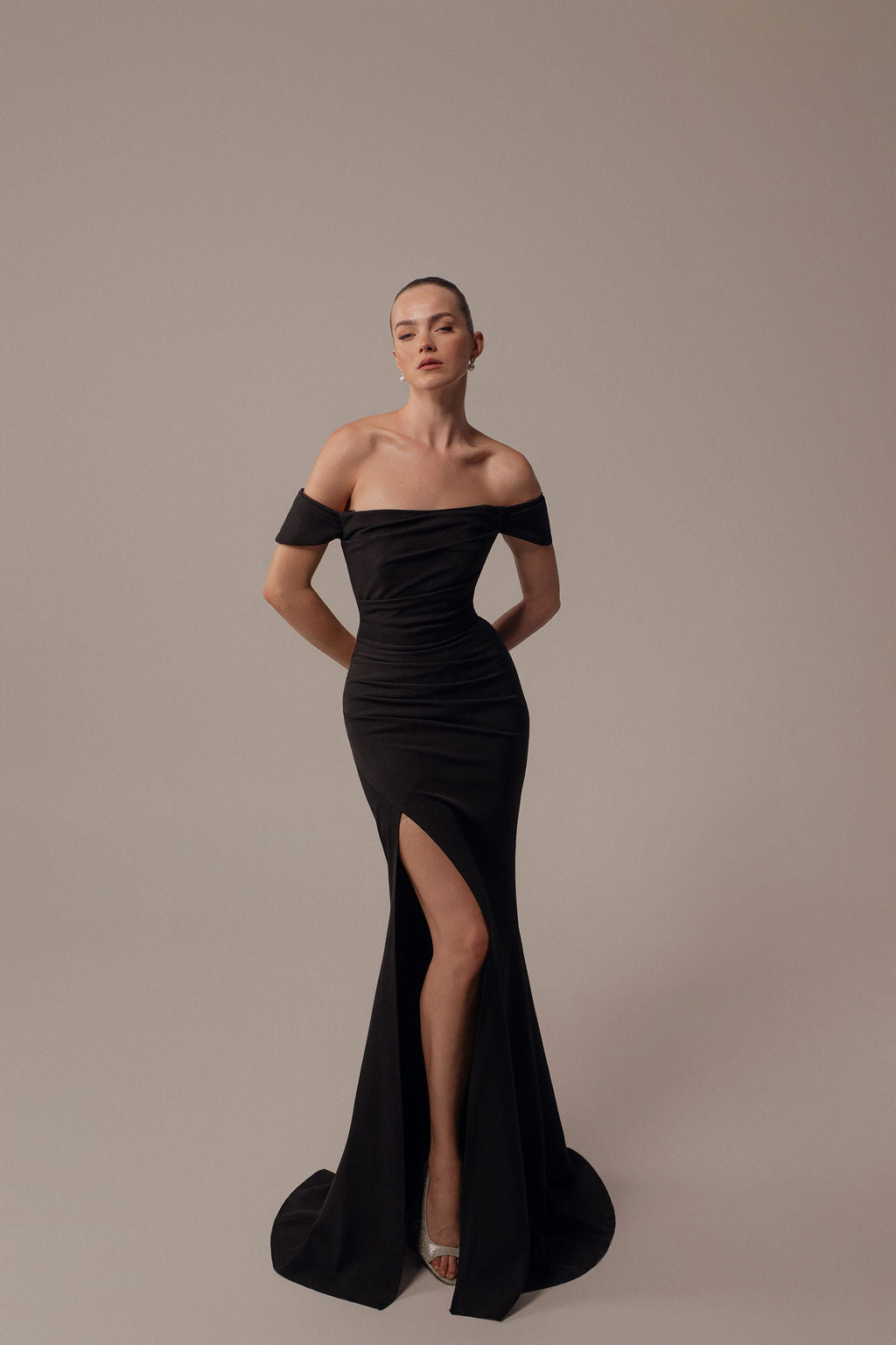אישה מדגמנת את שמלת Rona מקולקציית ערב 2026 של דרור קונטנטו — שמלת ערב שחורה צמודה עם כתפיים חשופות וחתך גבוה, כשהיא עומדת בביטחון על רקע בהיר, שיערה אסוף לאחור וזרועותיה מאחורי גבה.