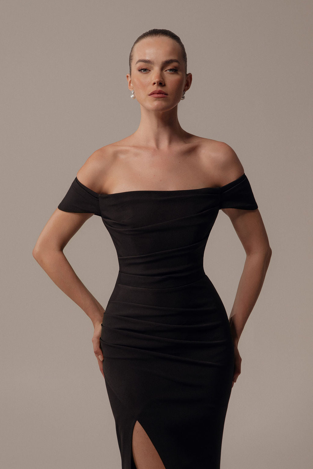 אישה מצטלמת בביטחון בשמלת Rona מקולקציית הערב 2026 של דרור קונטנטו — שמלת ערב שחורה צמודה עם כתפיים חשופות וחתך עד הירך. היא עומדת על רקע בז' ומשלבת את השמלה עם עגילי פנינה.