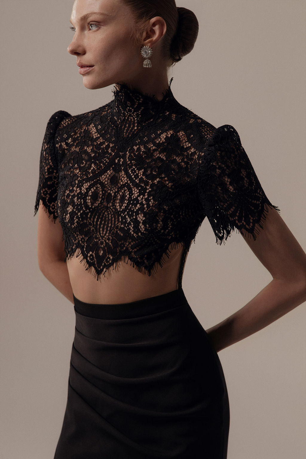 אישה לובשת את שמלת Hila מקולקציית ערב 2026 של דרור קונטנטו, הכוללת חולצת בטן שחורה עם צווארון גבוה וחצאית בגזרה גבוהה. שיערה מסודר לקוקו נמוך והיא עונדת עגילים בולטים, על רקע ניטרלי.