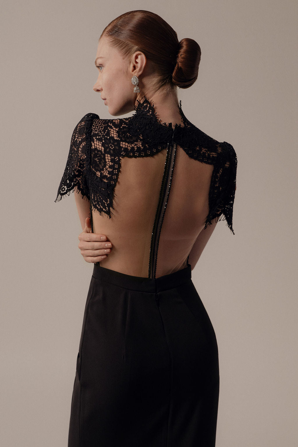 אישה עם שיער חום אסוף בקוקו חלק מדגמנת את שמלת Hila מקולקציית ערב 2026 של דרור קונטנטו, עם שרוולי תחרה קצרים וגב פתוח עם רוכסן גלוי. היא עומדת בצד, מביטה הצידה ומחזיקה את זרועה.