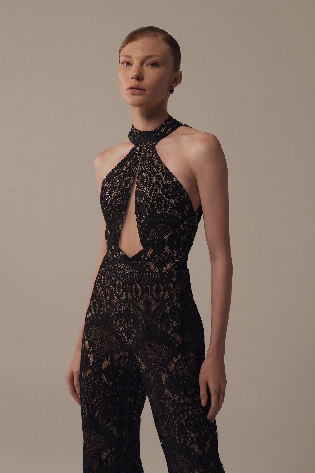 אישה בעלת עור בהיר ושיער חלק אסוף מדגמנת את בגד הגוף Gal - Jumpsuit מקולקציית Evening 2026 — בגד גוף שחור, ללא שרוולים, עם צווארון גבוה וחתך בחזה — על רקע ניטרלי וחלקה.