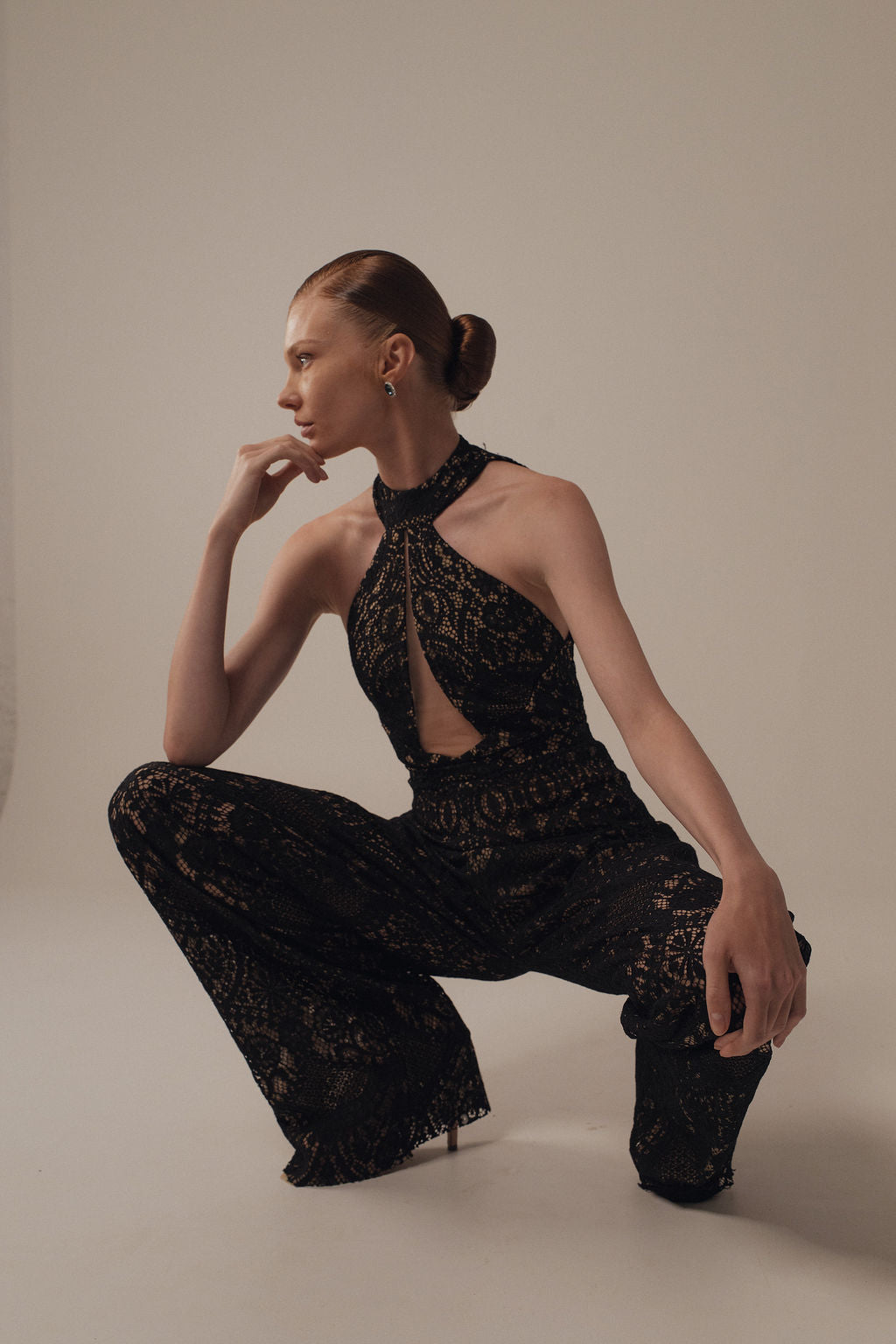 אישה עם שיער אסוף לאחור מדגמנת את בגד הגוף השחור ללא שרוולים Gal - Jumpsuit מקולקציית ערב 2026 של דרור קונטנטו, עם מחשוף סטרפלס וחתך קדמי, כורעת על ברך אחת על רקע בהיר וחלק.