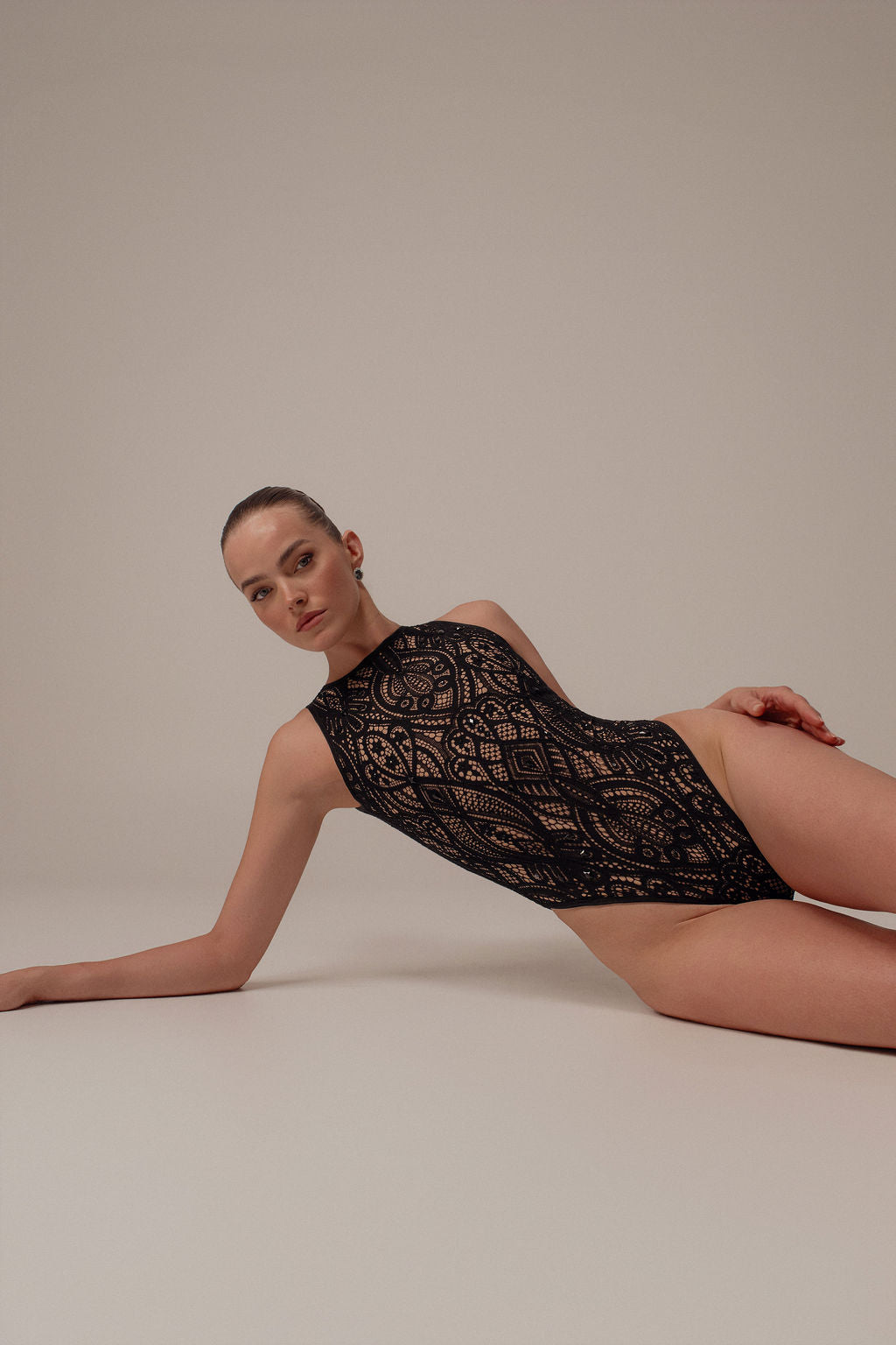 אישה נשענת על רקע חלק בבגד גוף שחור Inbar מקולקציית Evening 2026. שיערה חלק ומסורק לאחור, והבעת פניה ניטרלית, מה שמדגיש את העיצוב עם הצווארון הגבוה והשרוולים הקצרים.