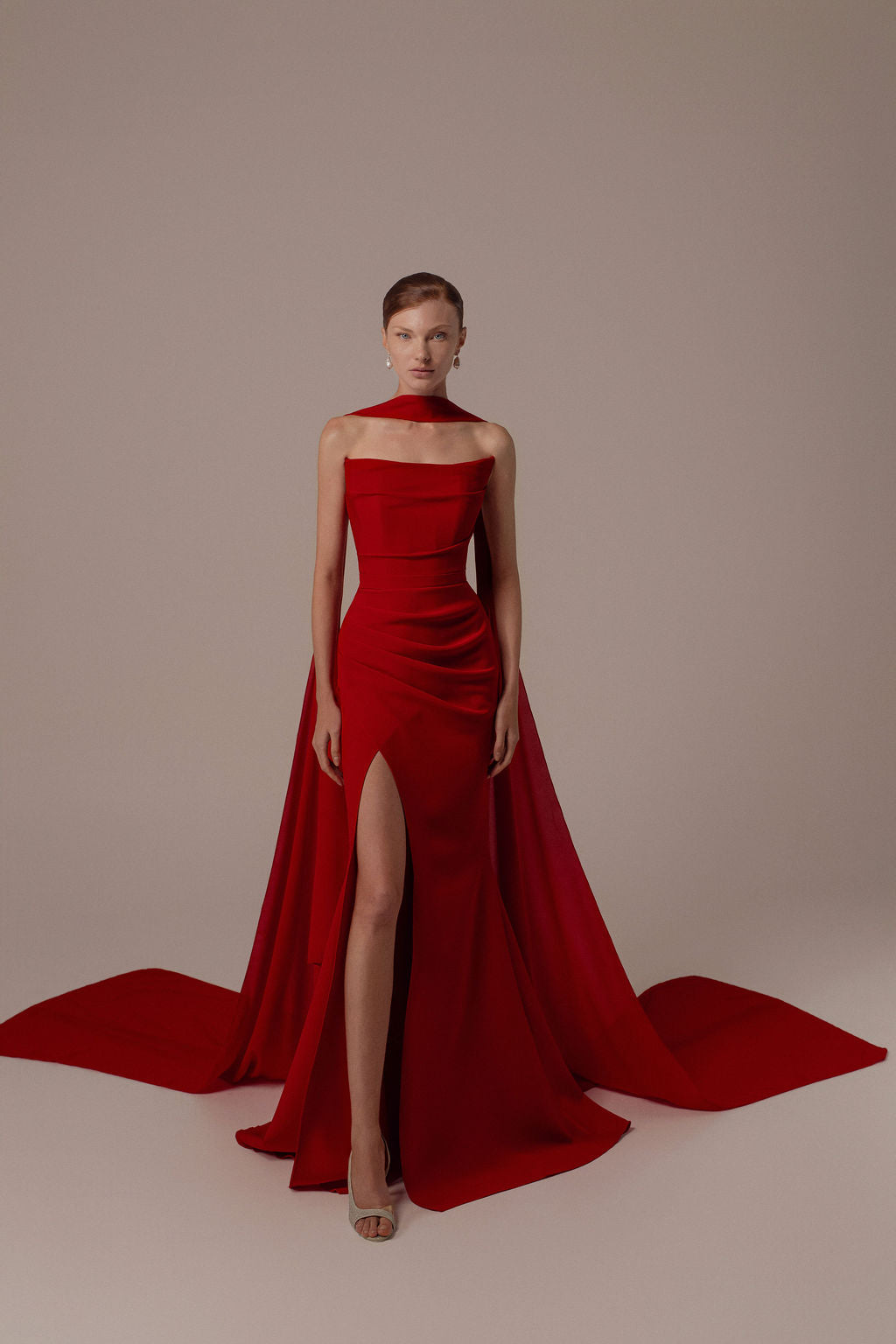 אישה לבושה בשמלת Tessa מקולקציית ערב 2026 בצבע אדום מבית Dror Kontento עומדת על רקע בהיר. השמלה ללא כתפיות כוללת שובל דרמטי, חתך גבוה, תכשיטים מינימליים ושיער אסוף לאחור למראה אלגנטי.