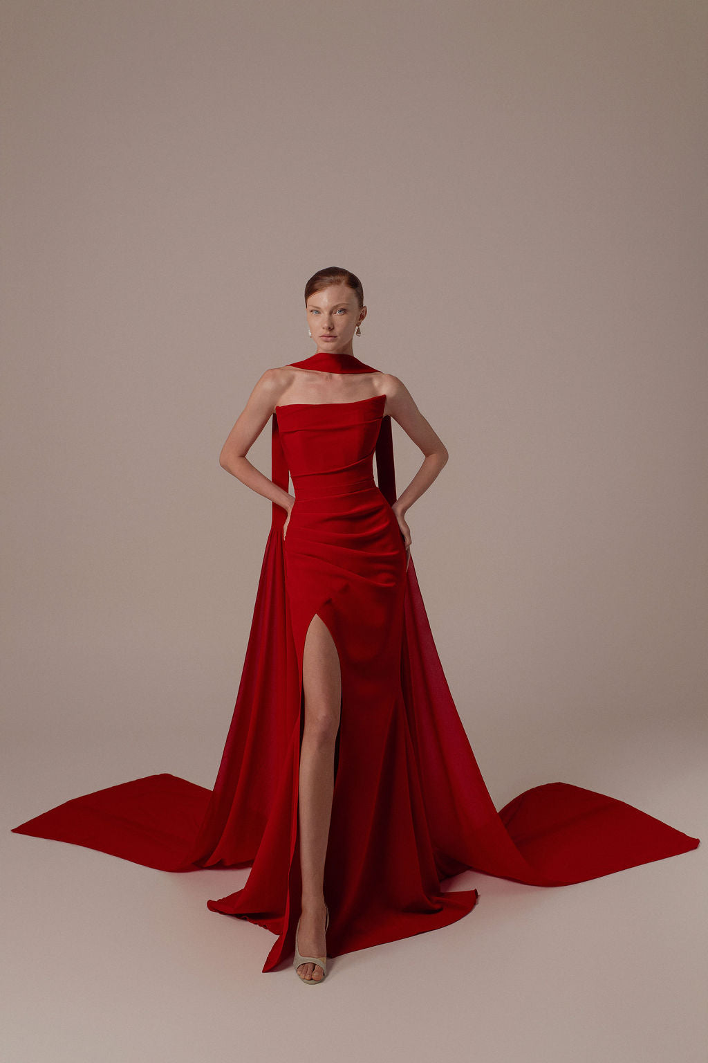 אישה עומדת בביטחון על רקע ניטרלי, ידיה על מותניה, לבושה בשמלת Tessa מקולקציית הערב 2026 של דרור קונטנטו — שמלת ערב אדומה בולטת עם חתך גבוה ושובל ארוך וזורם. היא מביטה ישירות למצלמה.