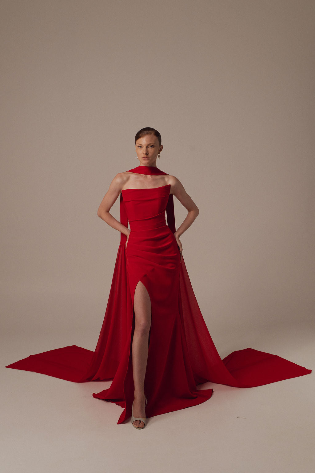 אישה מצטלמת בביטחון על רקע בהיר, ומציגה את שמלת Miri מקולקציית ערב 2026 — שמלת ערב אדומה ומרשימה ללא כתפיות, עם חתך גבוה עד הירך ושובל ארוך וזורם.