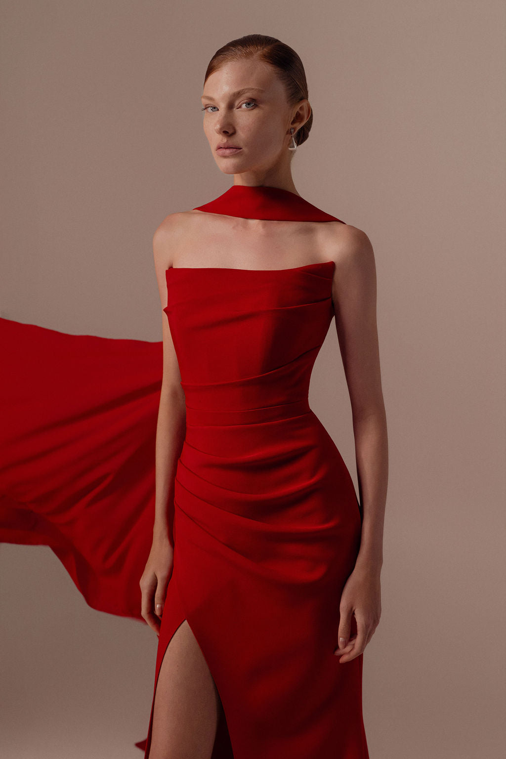 אישה לבושה בשמלת Tessa מקולקציית ערב 2026 של דרור קונטנטו, עם גזרה צמודה ללא כתפיות, מחשוף א-סימטרי וחתך גבוה, מצטלמת על רקע ניטרלי עם צעיף אדום תואם.