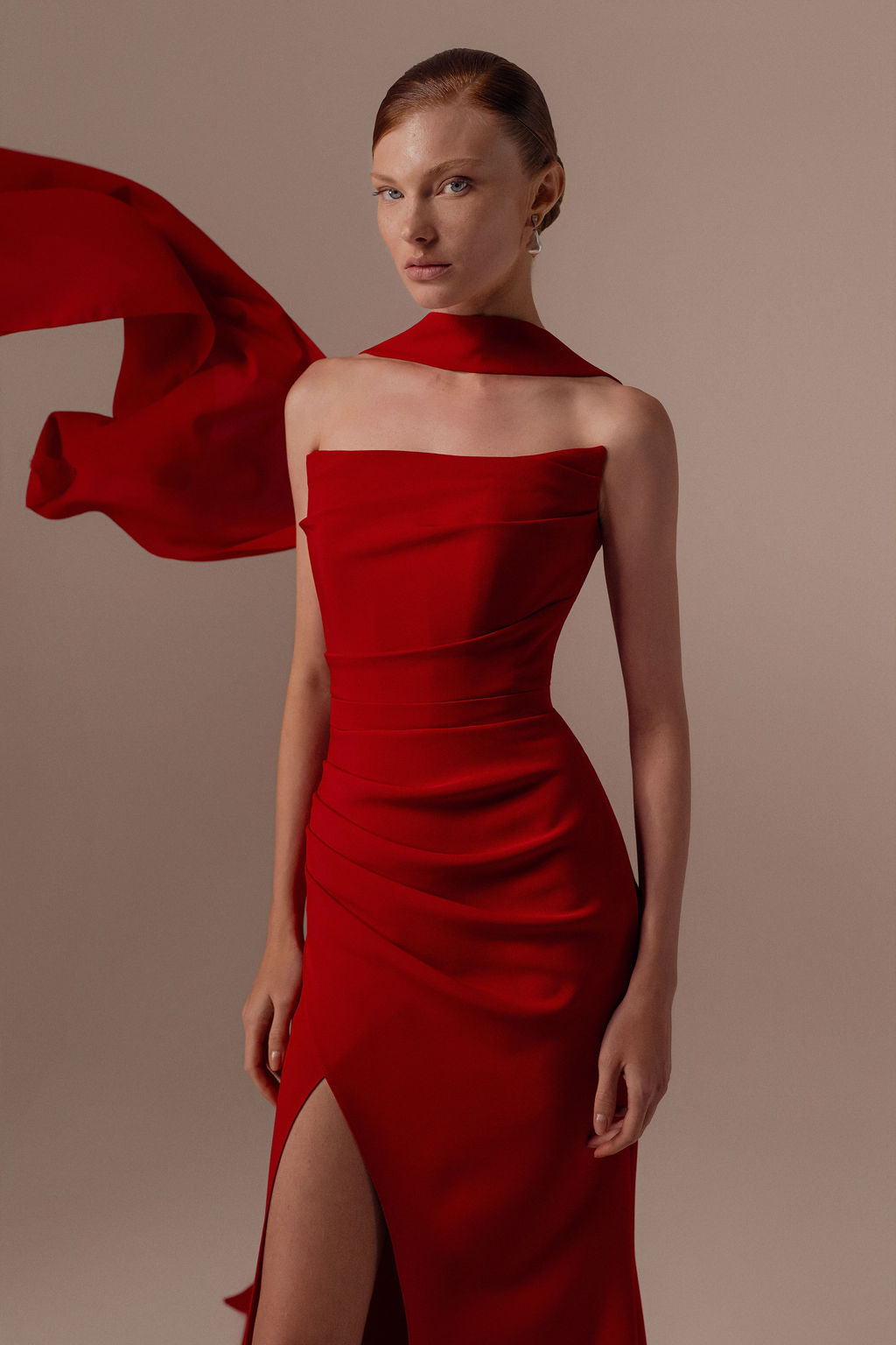 אישה לבושה בשמלת Tessa - קולקציית ערב 2026 אדום מאת דרור קונטנטו, שמלת סטרפלס אלגנטית עם חתך גבוה וצעיף אדום תואם, עומדת על רקע ניטרלי ומביטה בביטחון למצלמה.