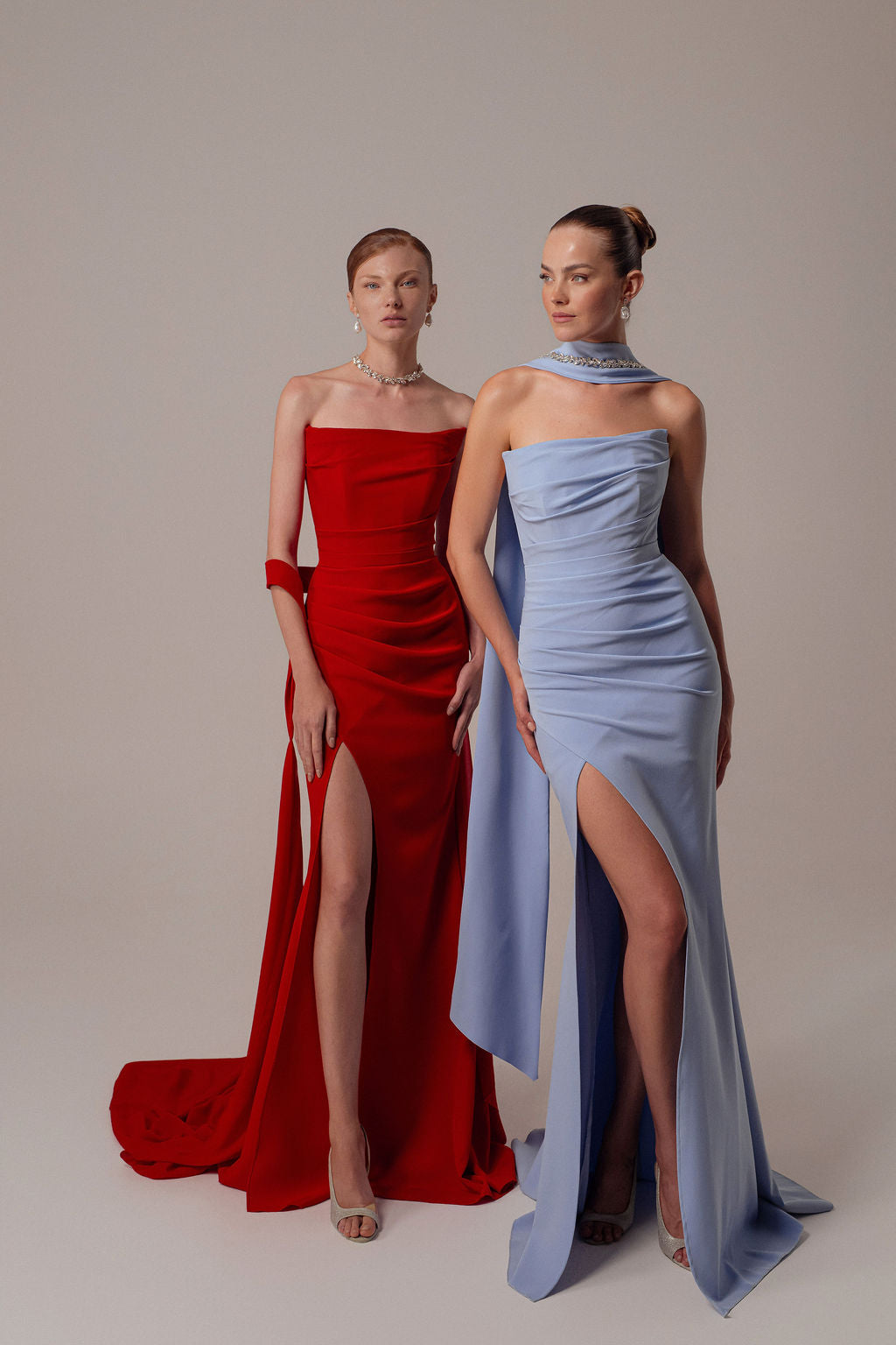 שתי נשים עומדות זו לצד זו בשמלות ערב אלגנטיות — האחת לובשת את שמלת Tessa מקולקציית Evening 2026, שמלת ערב אדומה ללא כתפיות של דרור קונטנטו, והשנייה לובשת שמלה סטרפלס בצבע תכלת. שתיהן עם תסרוקות אסופות, נעלי עקב ושרשראות בולטות.