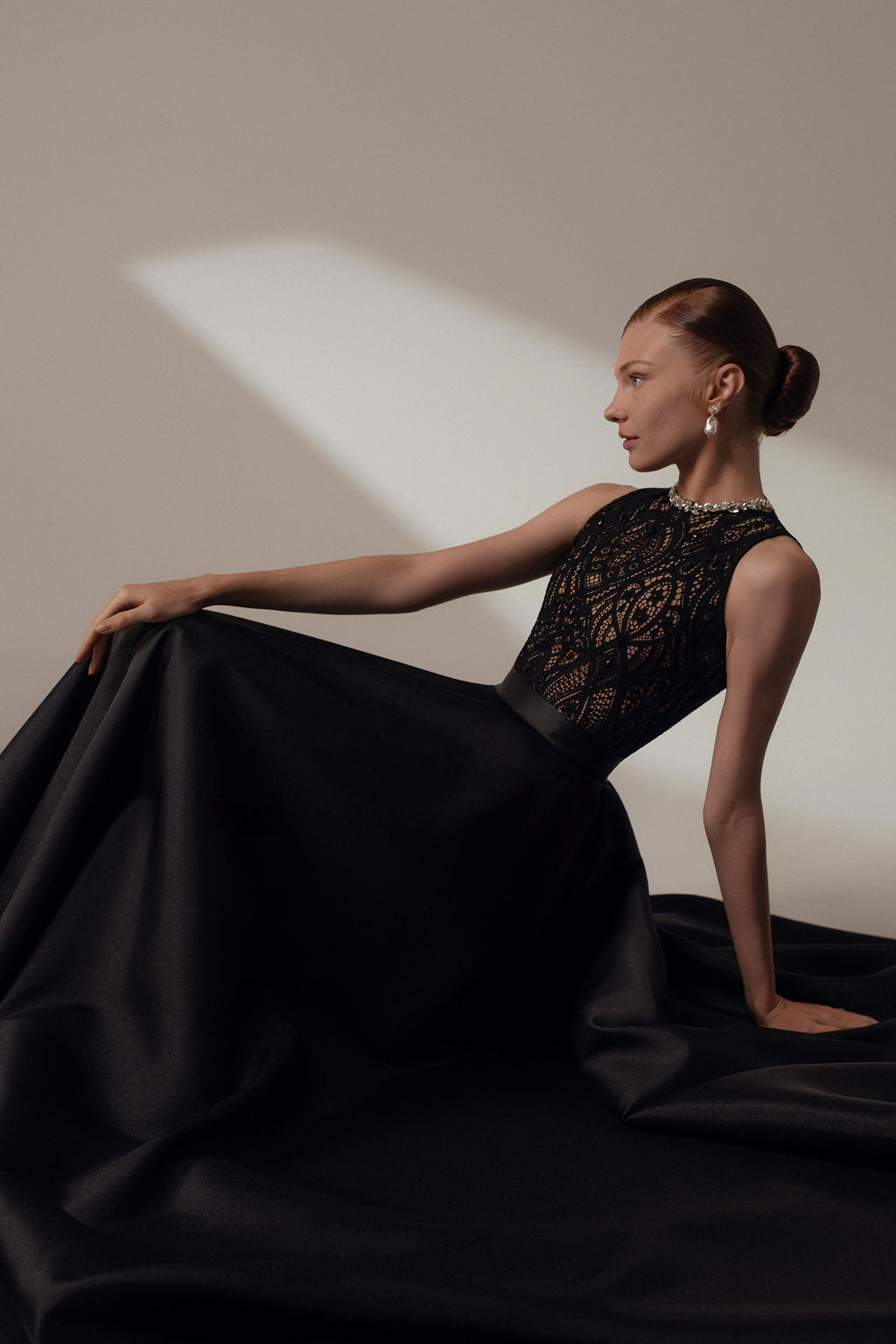 אישה לבושה בשמלת Lia מקולקציית ערב 2026 של דרור קונטנטו, עם מחוך תחרה וחצאית מלאה, יושבת בחינניות על הרצפה. התסרוקת האלגנטית והעגילים הארוכים משלימים את הרקע הנייטרלי.