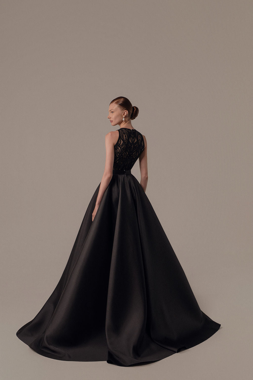 אישה עומדת בחינניות בשמלת Lia מקולקציית ערב 2026 של דרור קונטנטו, עם חצאית מלאה ופרטי תחרה בגב. שיערה אסוף בקוקו חלק והיא פונה עם גבה למצלמה בסטודיו ניטרלי.