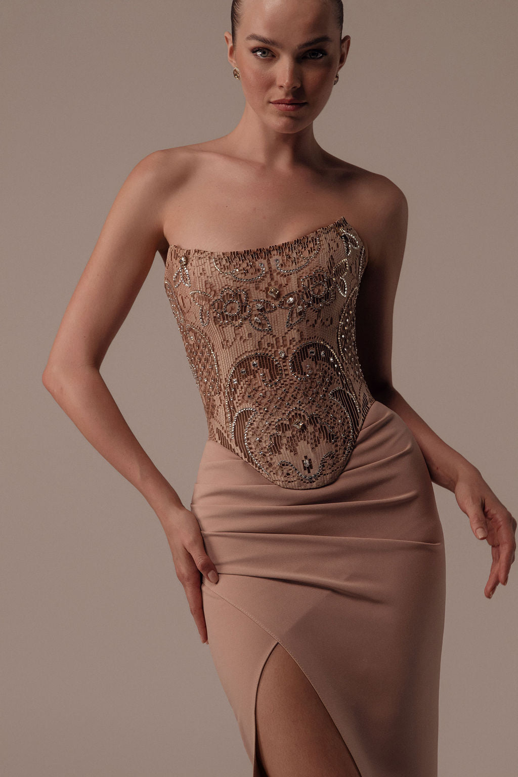 אישה מדגמנת את שמלת Dana מקולקציית ערב 2026 של דרור קונטנטו, עם גזרה מחמיאה ללא כתפיות, מחוך מעוטר בחרוזים וחתך גבוה בחצאית על רקע בז' חלק.